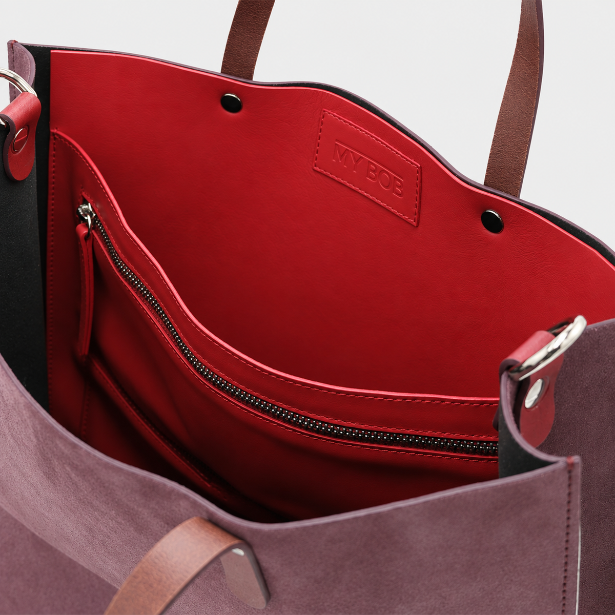 Close-up of the interior of a tote bag showing a bright red lining with a zippered pocket and a small embossed MY BOB brand patch. The bag also has mauve-purple exterior suede leather and brown handles.