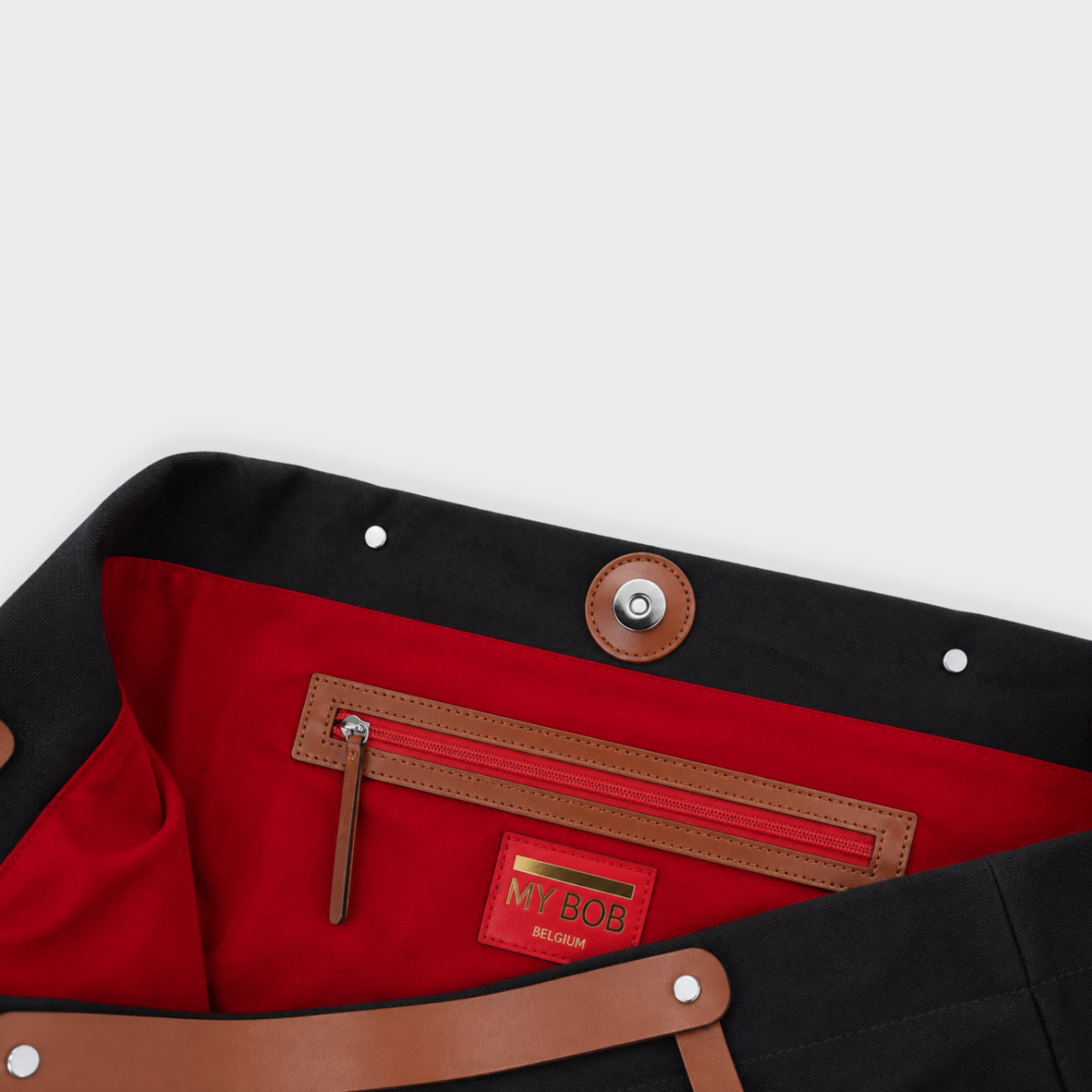 Close-up detail of a black canvas bag, showing a bright red interior lining with a brown leather zipper pocket and a red brand patch reading 'MY BOB'.