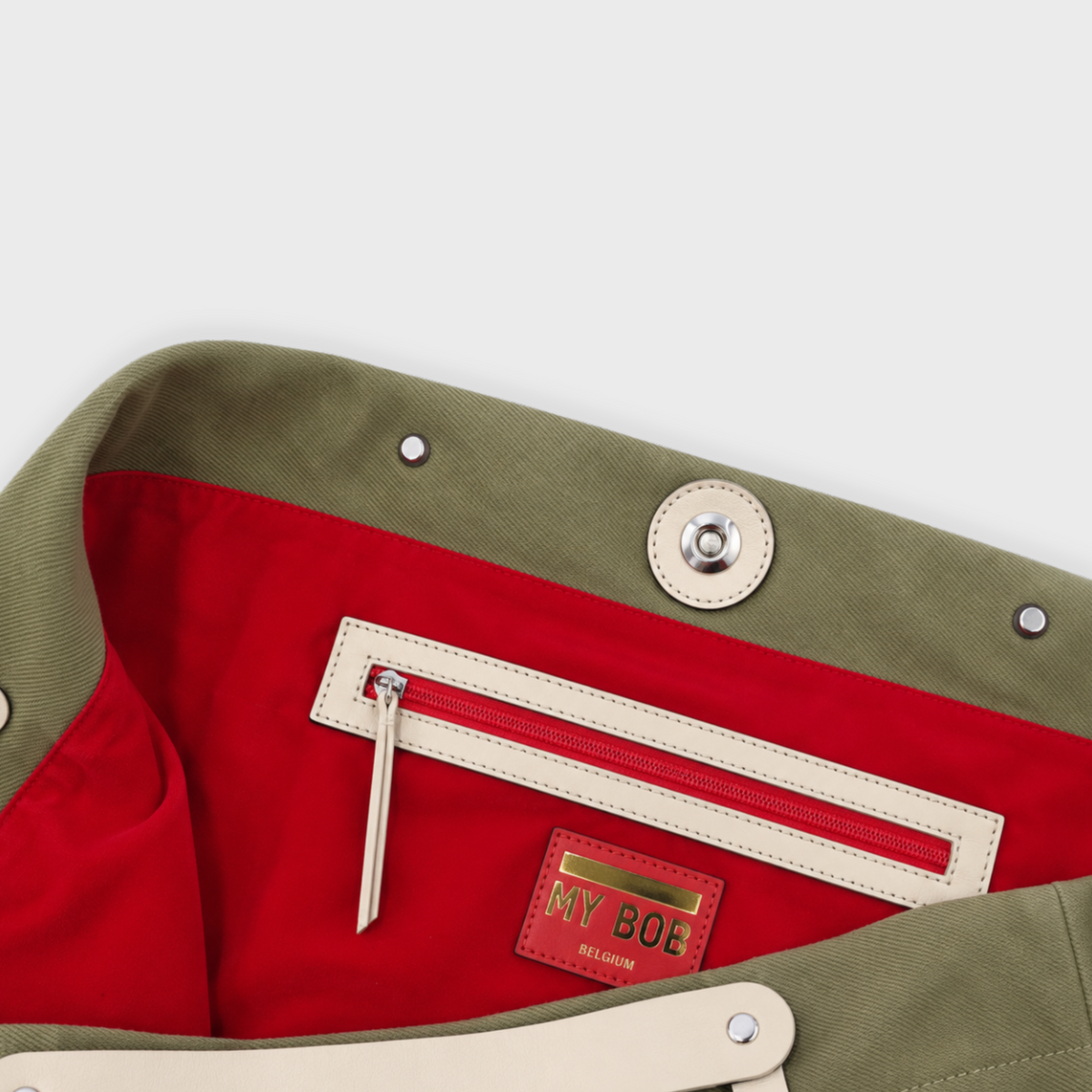 Close-up of a canvas bag interior showing an olive green exterior, a bright red lining, a cream zipper pocket, and a red leather patch with the brand name 'MY BOE'.