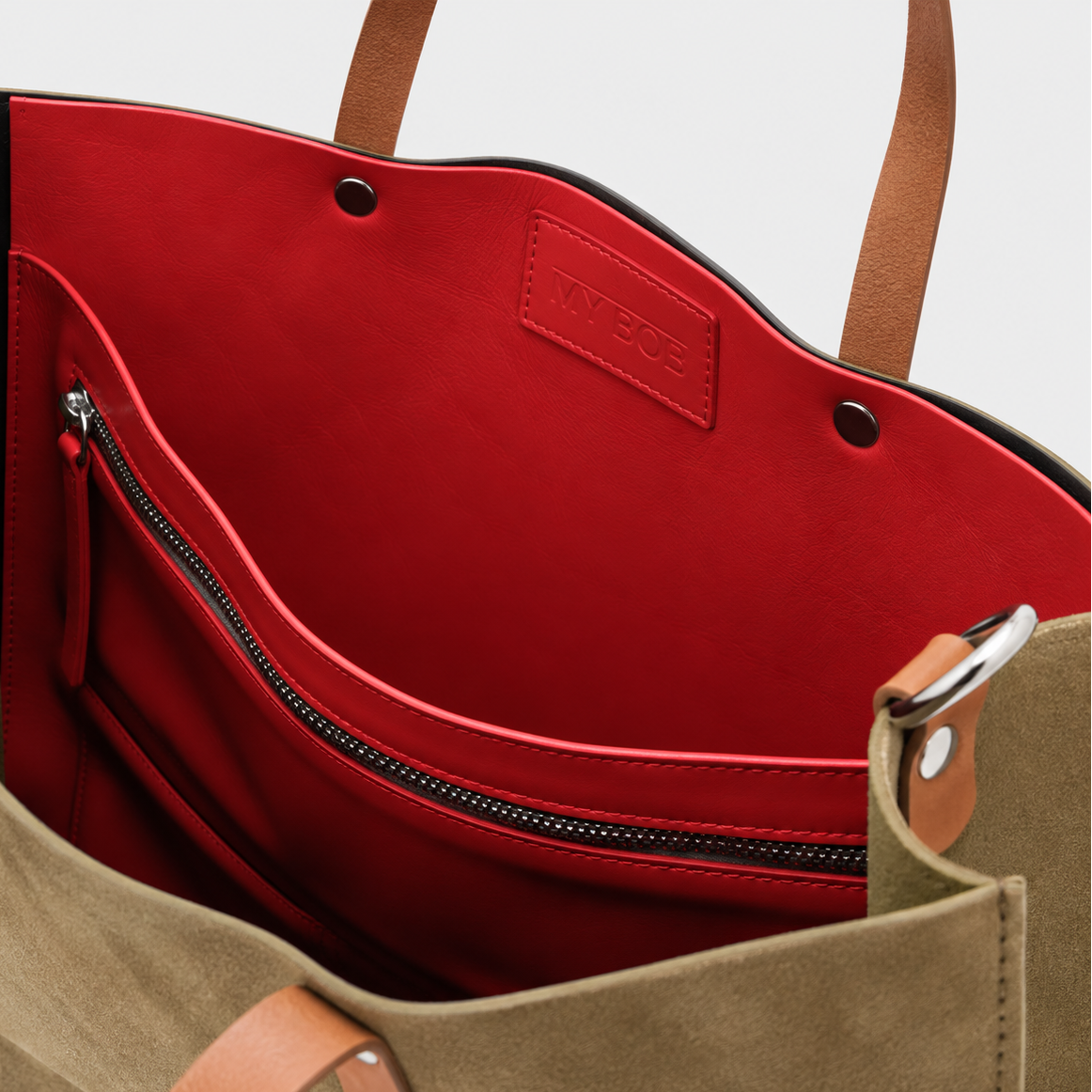 Close-up of a tote bag interior showing a bright red leather lining with a zippered pocket, tan leather handles, and a suede exterior in a muted olive-tan color.