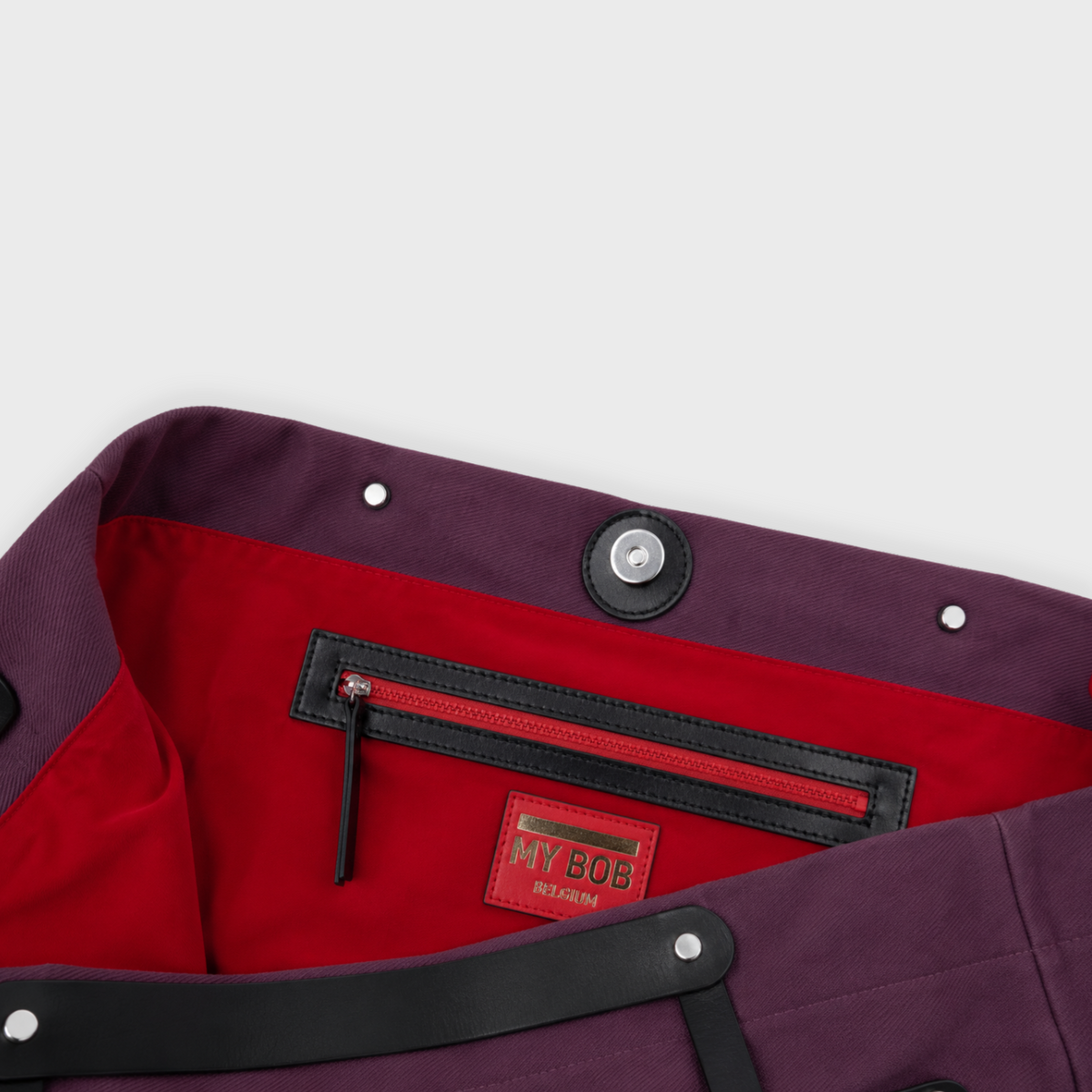 Close-up of a dark purple bag with a bright red interior lining, featuring a black zippered pocket and a red 'MY BOY' brand patch.