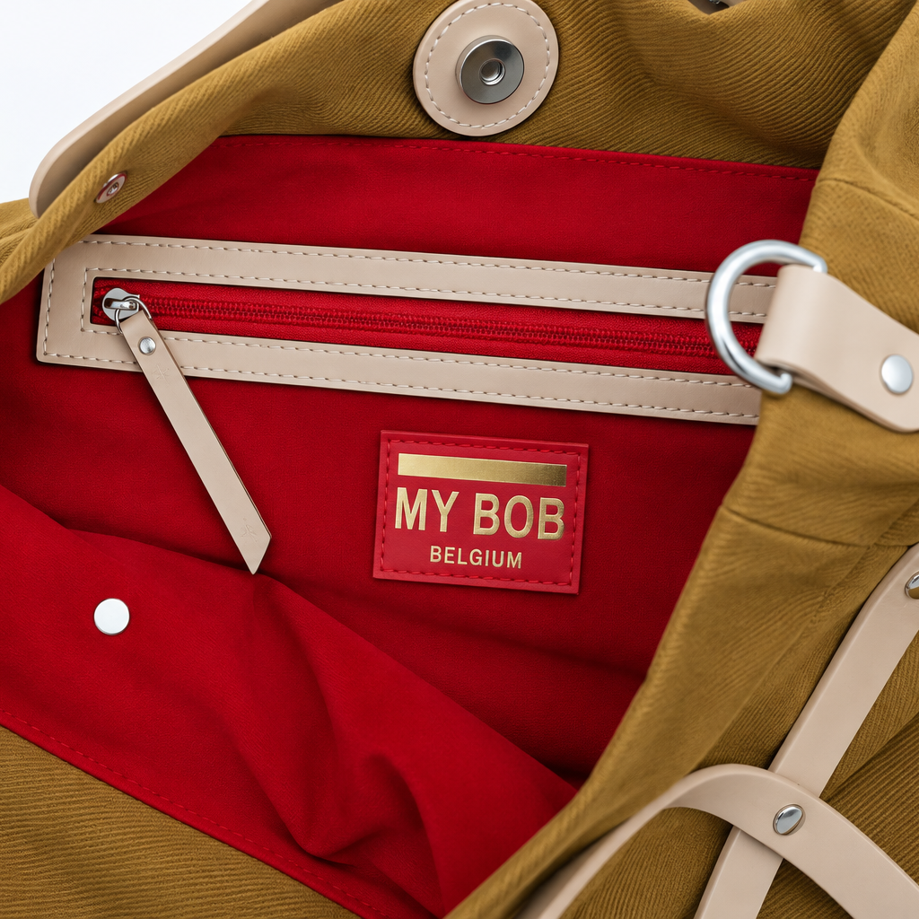 Close-up of a mustard yellow canvas bag with a bright red interior lining, beige leather trim, metal hardware, and a red patch label reading 'MY BOB BELGIUM'.