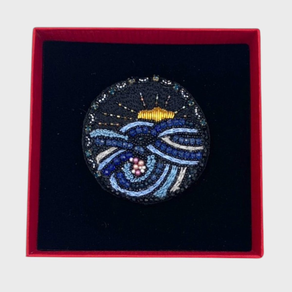 Decorative brooch with blue and gold embroideries in a red box on a white background