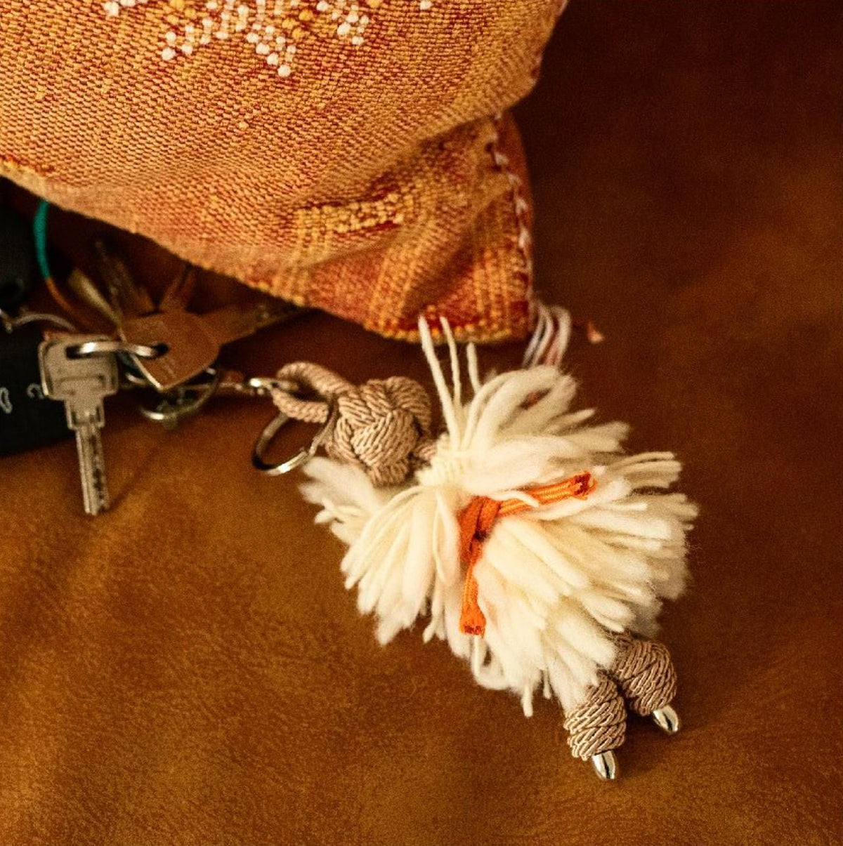 A beige and brown key ring with a macramé pattern, attached to a keychain, placed on a leather surface.