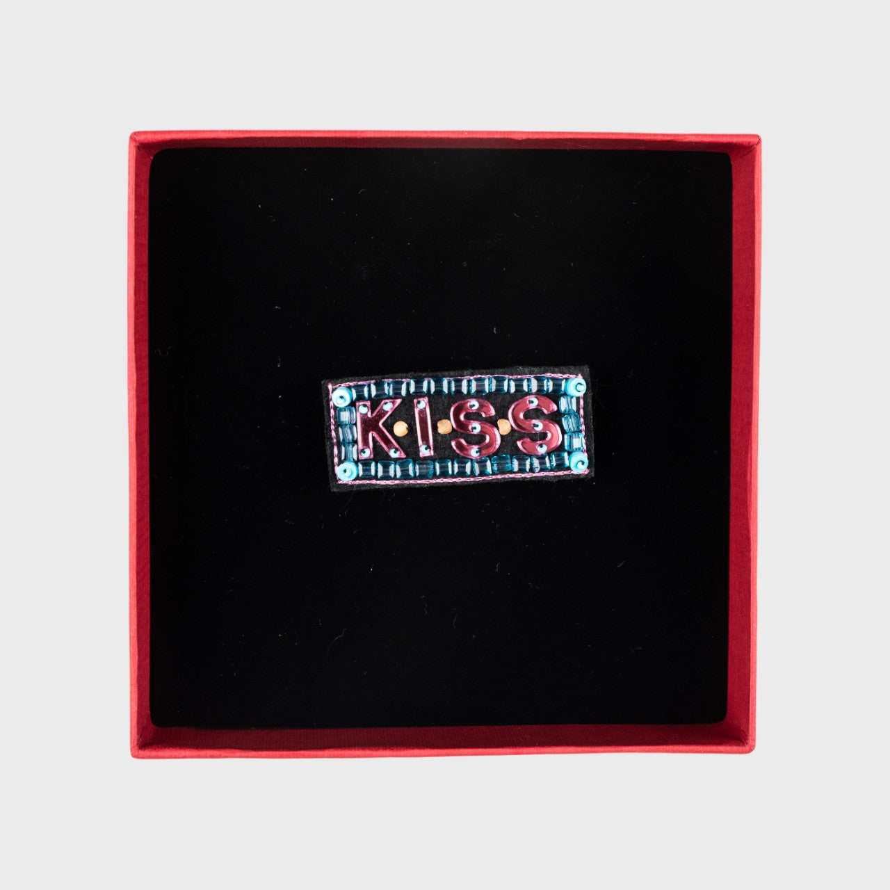 An embroidered KISS brooch with pink and blue details in a box. 