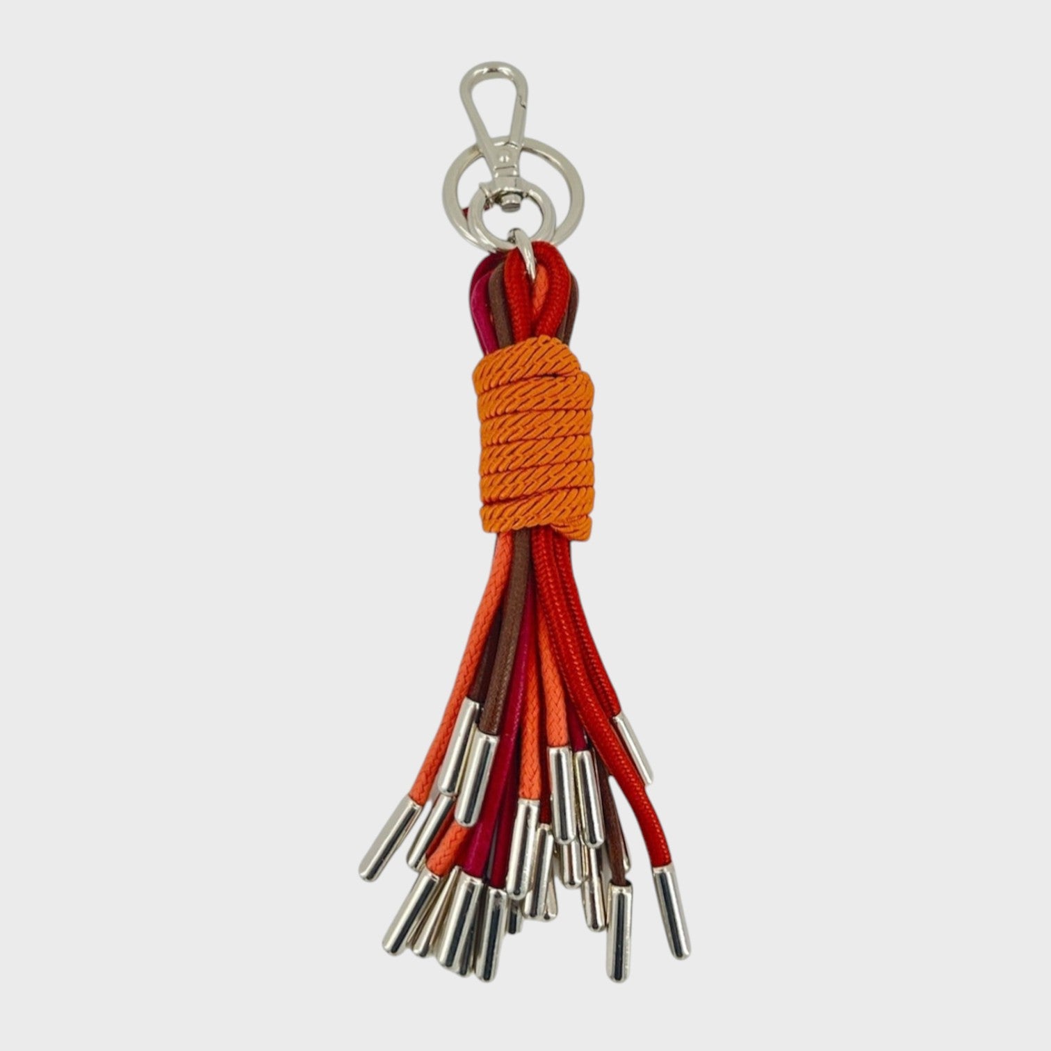 A keychain tassel with a silver metal clasp and ring, featuring an orange woven wrap and multiple hanging cords in red, brown, and orange.