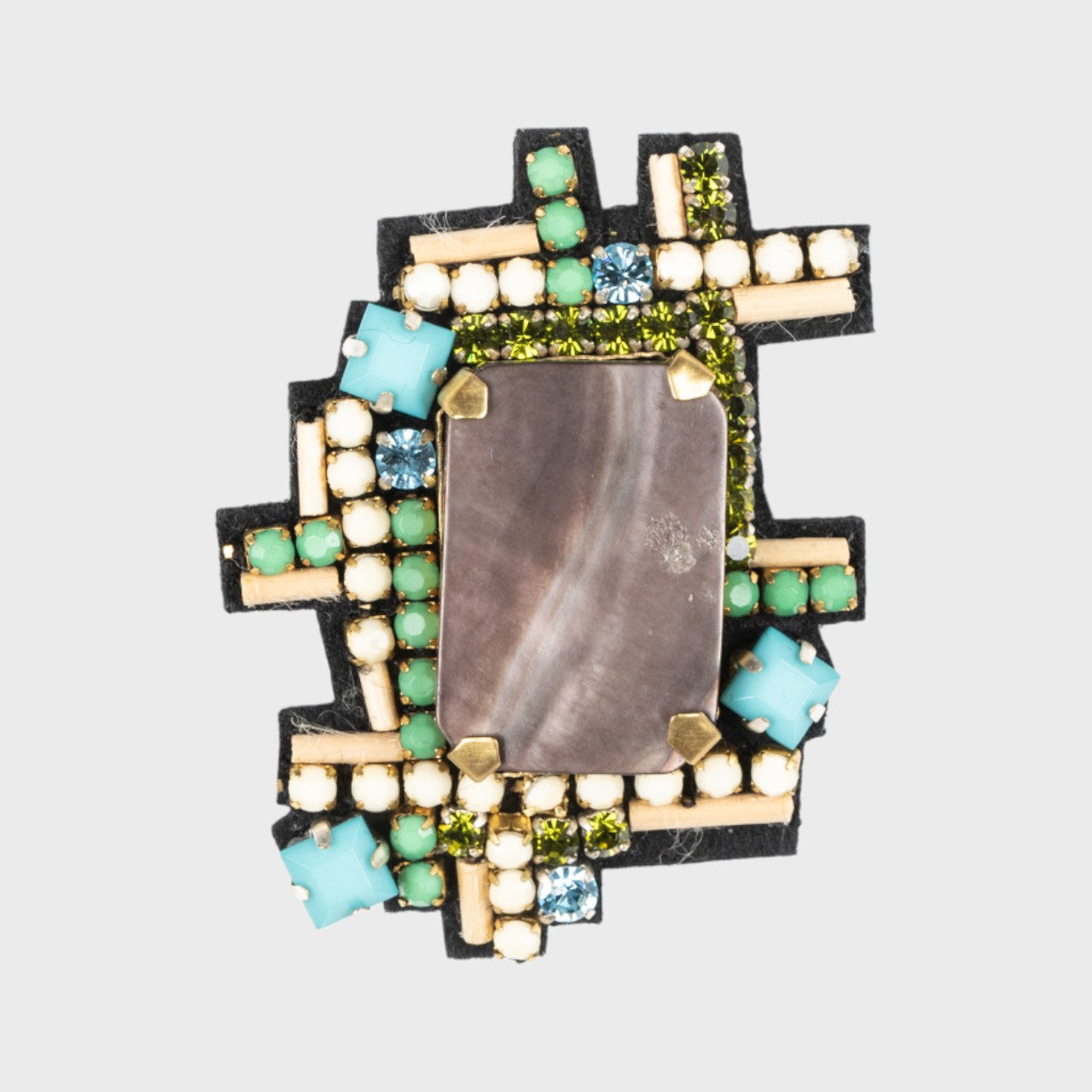 An embroidered brooch symbolizing a lake with a stone in the middle and green, blue and beige details around. 