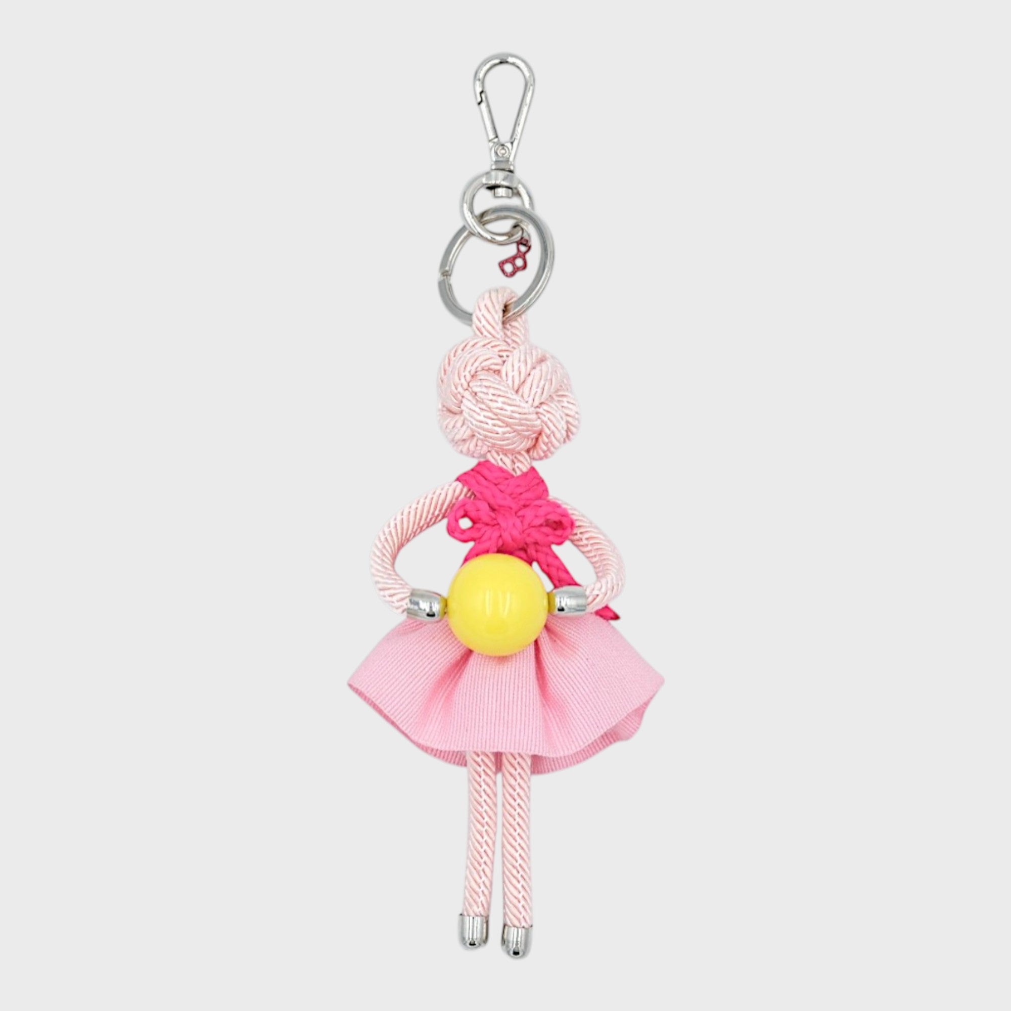 Handmade macramé bag charm featuring an all pink colored character in light pink skirt, a dark pink bow top, holding a yellow ball. She is attached to a silver clasp.