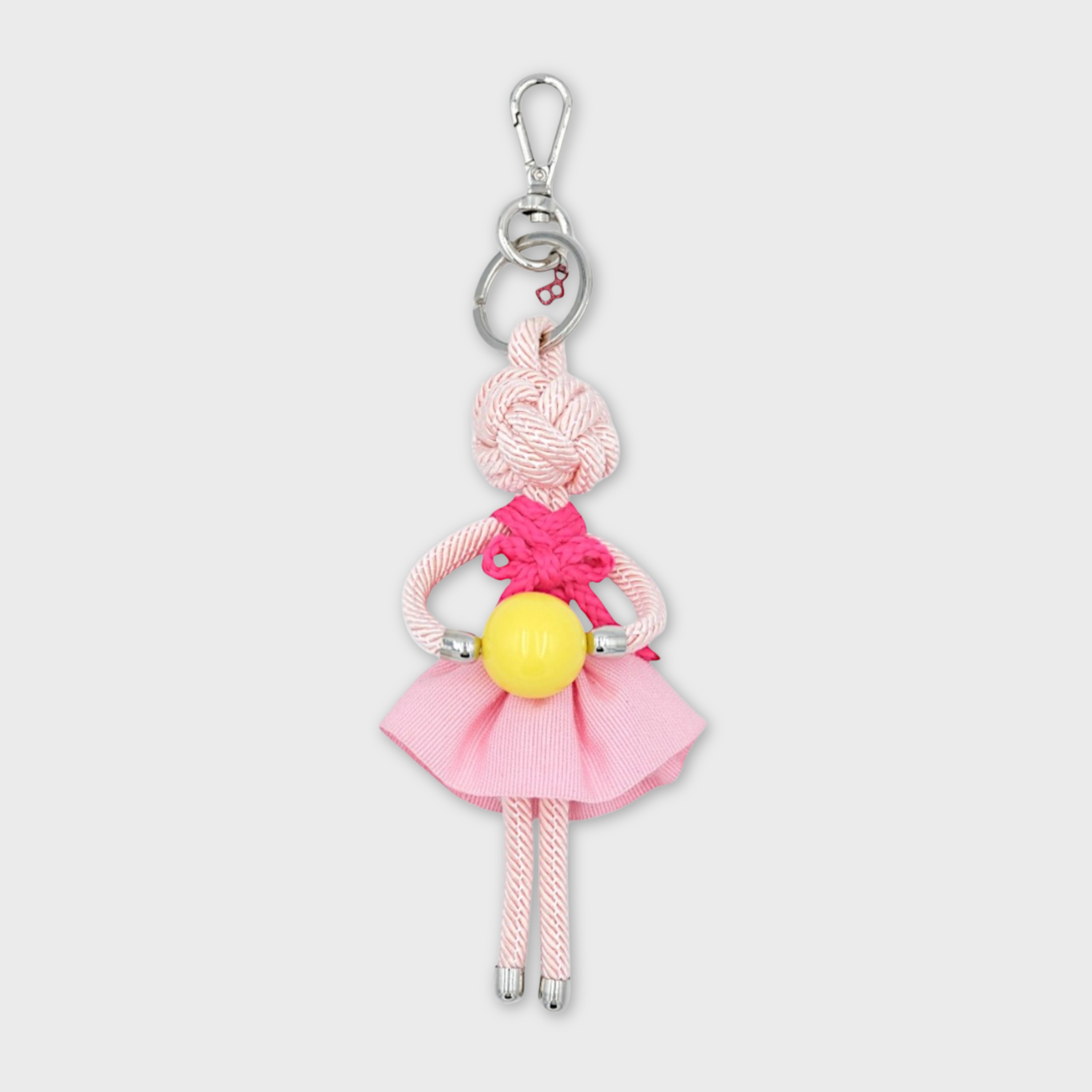 Handmade macramé bag charm featuring an all pink colored character in light pink skirt, a dark pink bow top, holding a yellow ball. She is attached to a silver clasp.