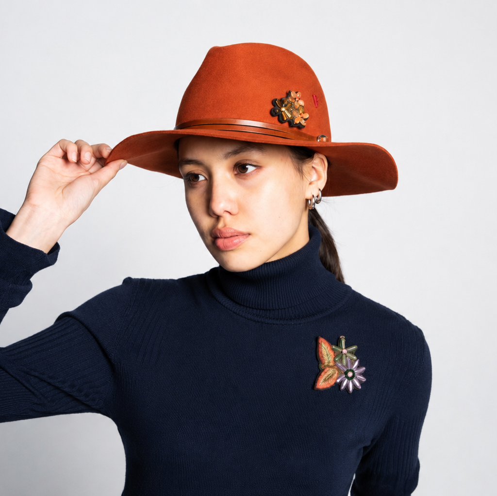 A woman wearing a rust-orange wide-brim fedora hat with a flower brooch and a dark navy ribbed turtleneck sweater with a matching flower chest brooch, posing against a light gray background.