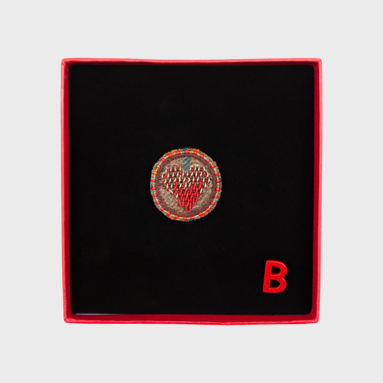 An embroidered brooch representing a red heart in the middle of a brown circle in a box. 