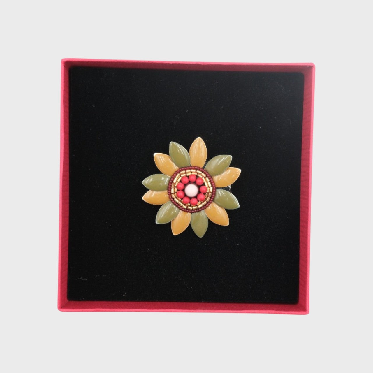 An embroidered brooch representing a green and beige manuka flower in a box. 