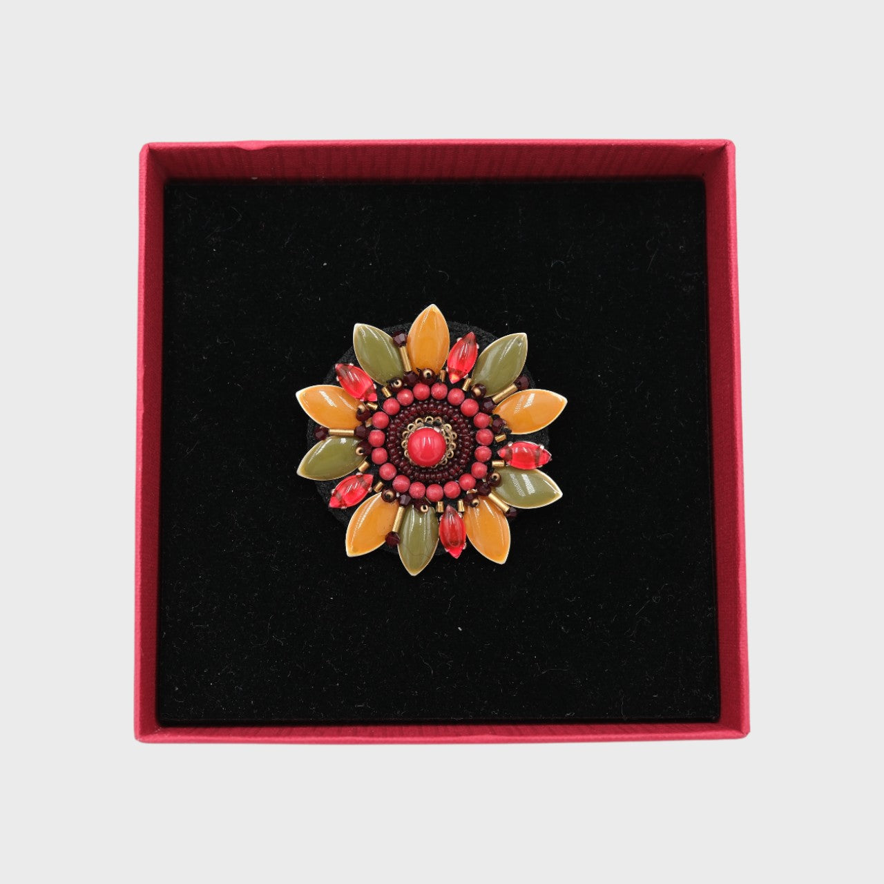 An embroidered brooch representing a manuka flower with red, green and beige details in a box. 
