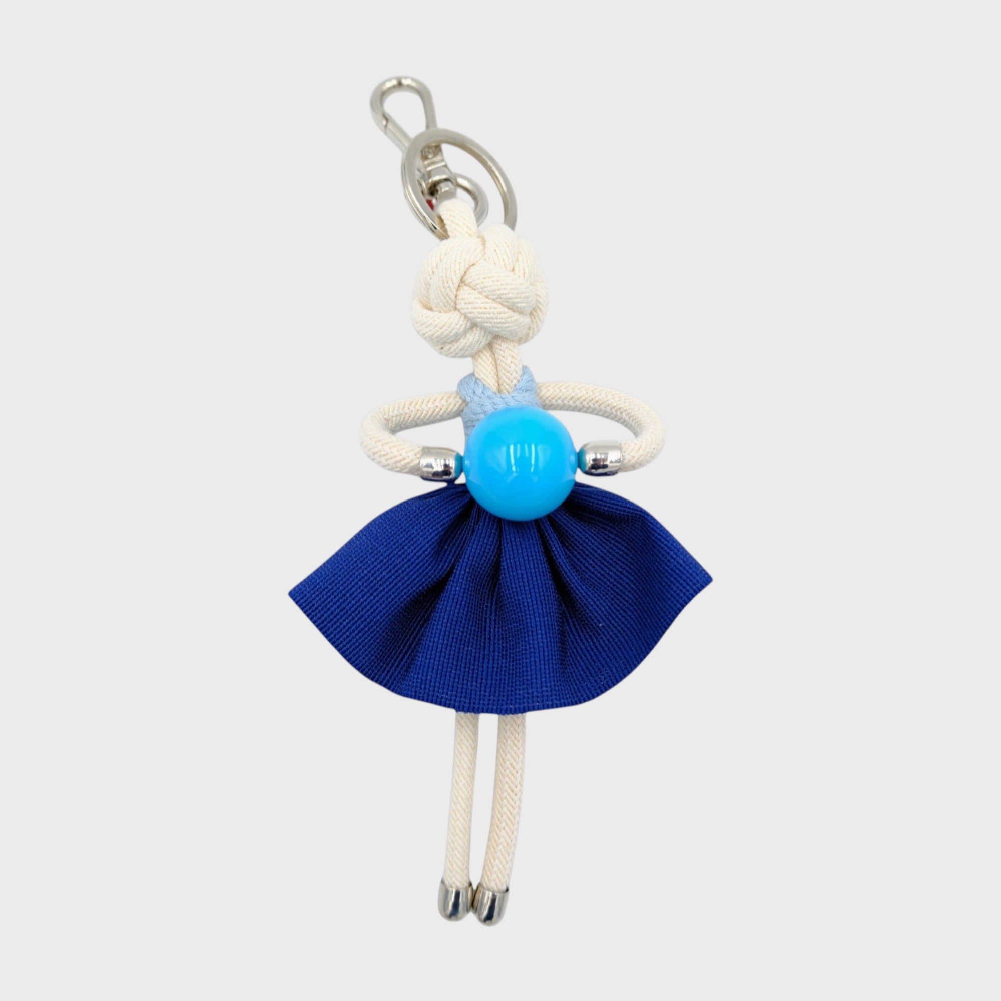 Handmade white macramé bag charm featuring a character in dark blue mini skirt, a light blue top, holding a light blue ball. She is attached to a silver clasp.