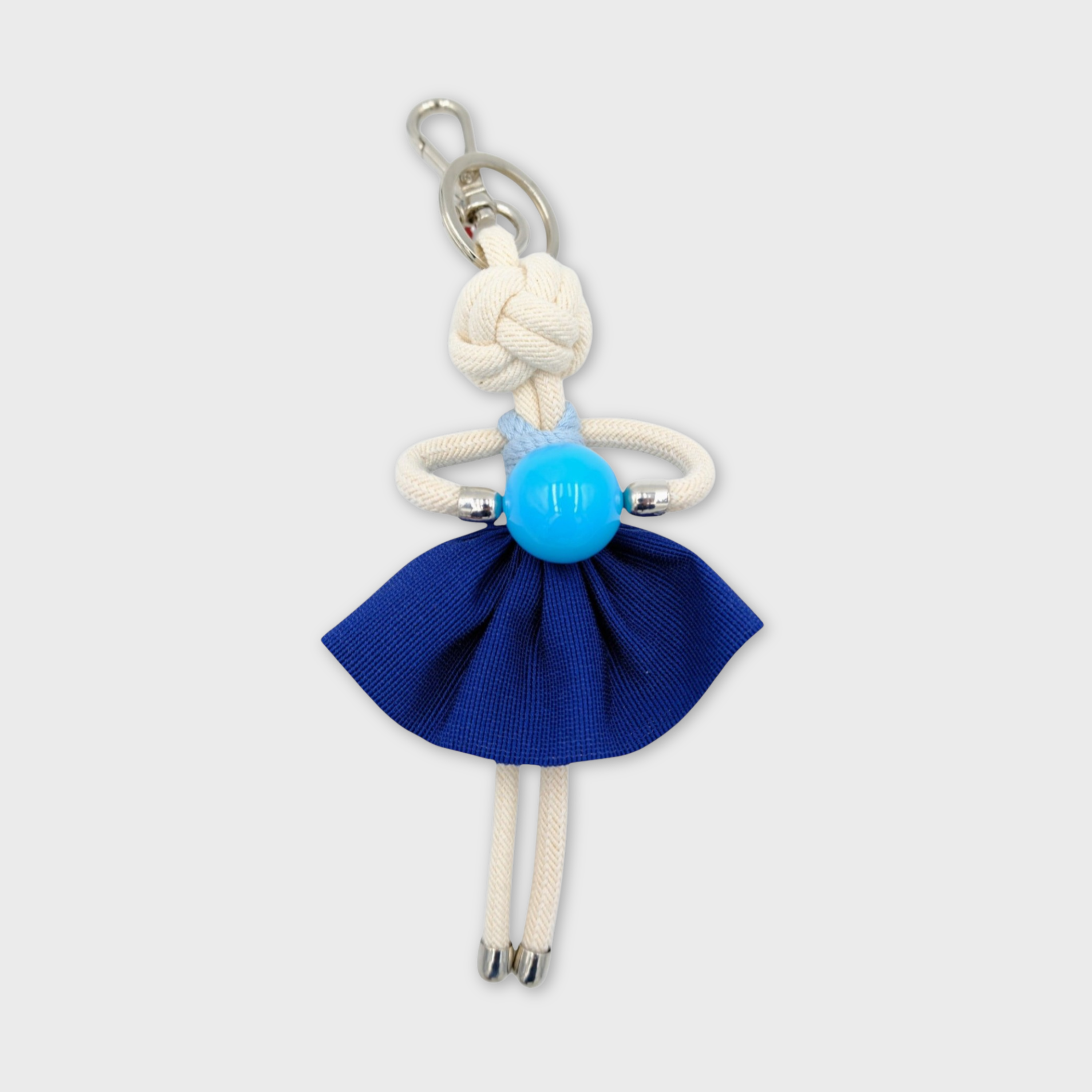 Handmade white macramé bag charm featuring a character in dark blue mini skirt, a light blue top, holding a light blue ball. She is attached to a silver clasp.