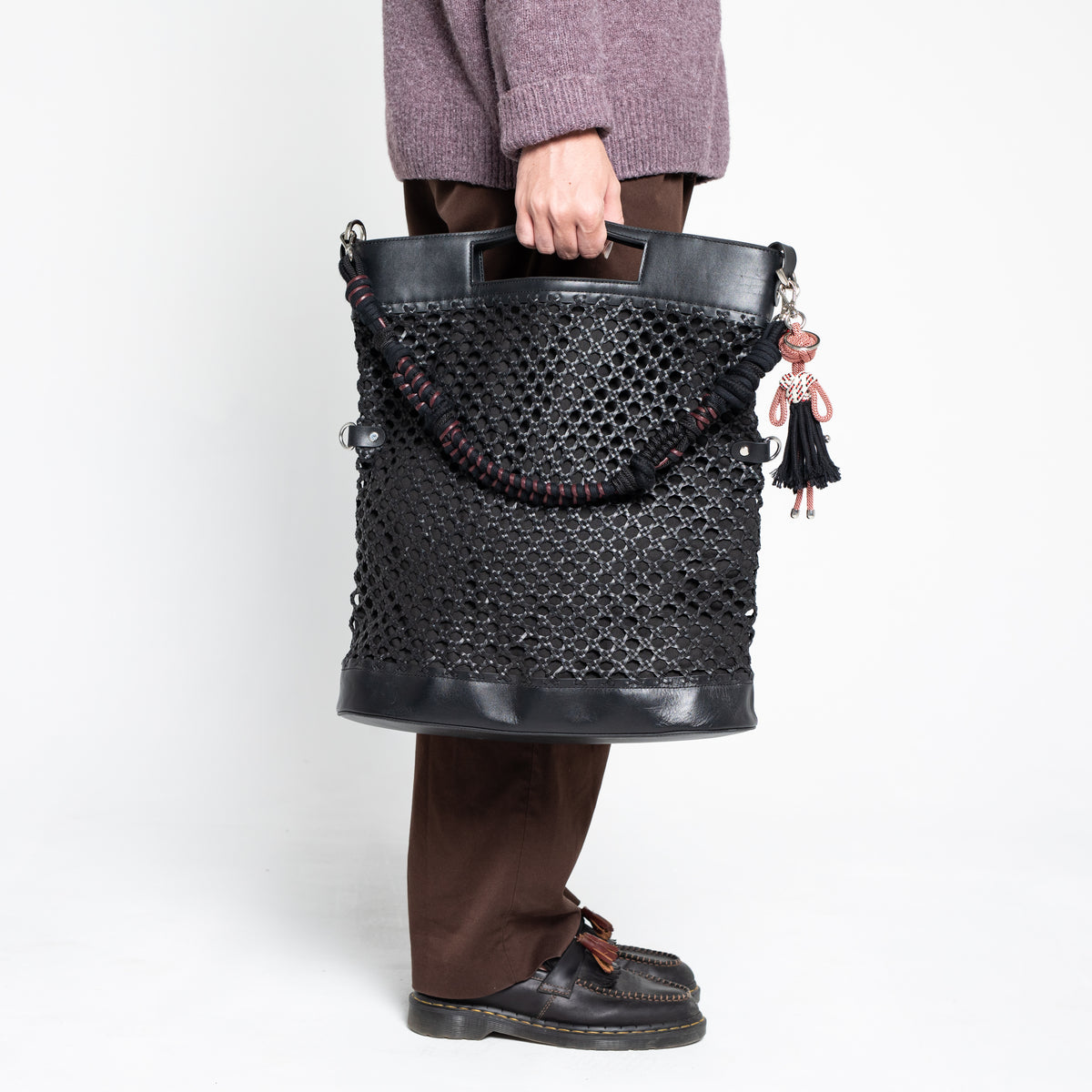Person holding a black textured handbag  with a red and brown macramé braided strap on a white background