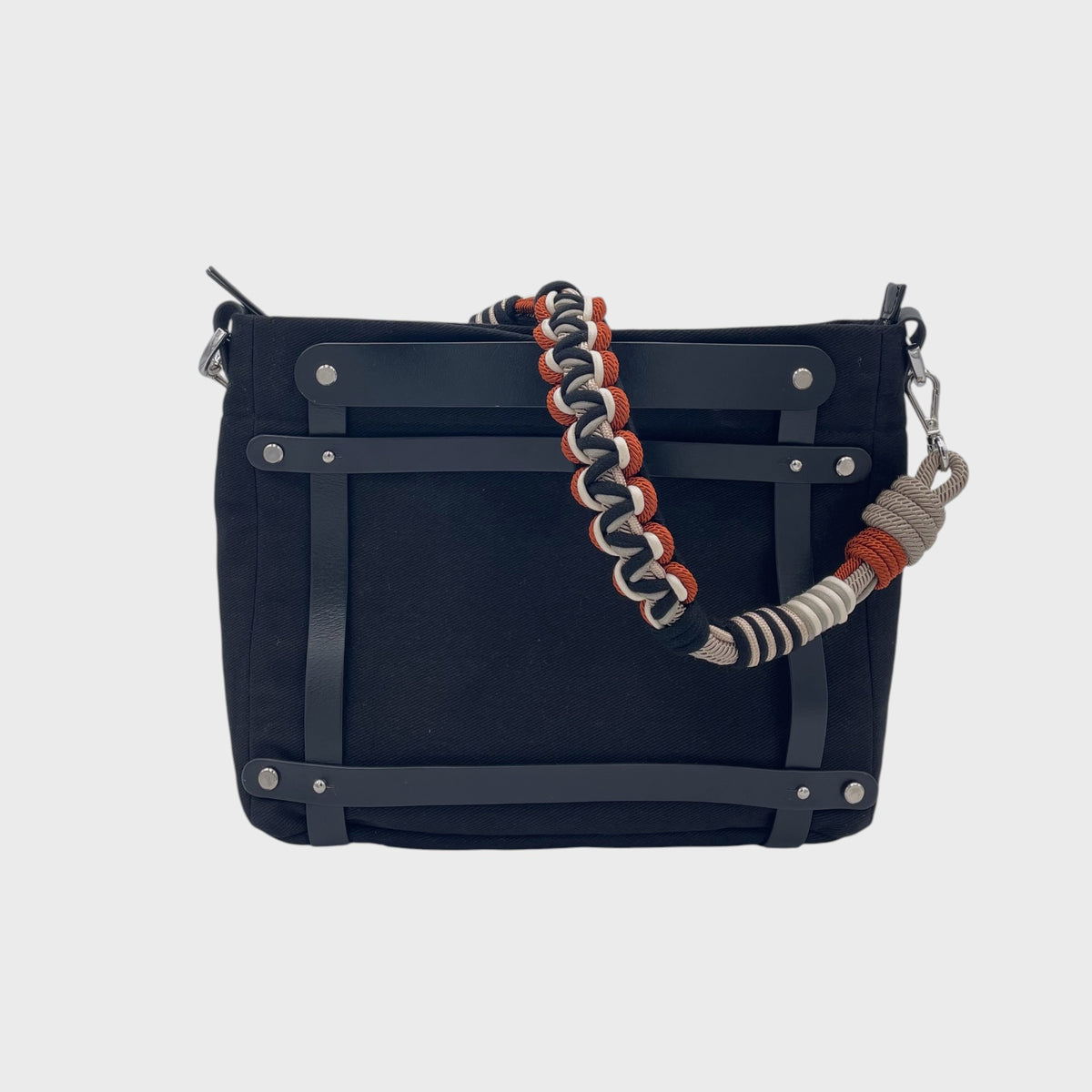 Black cotton canvas handbag with black leather harness and a braided multicolor macrame strap in red, black and grey on a light gray background.