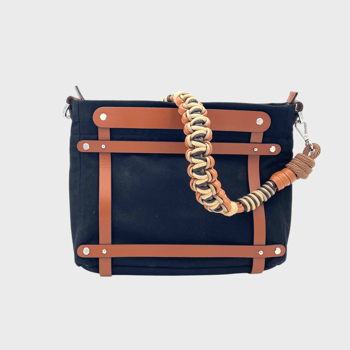 Black handbag with brown leather straps and a braided macrame handle on a light gray background.