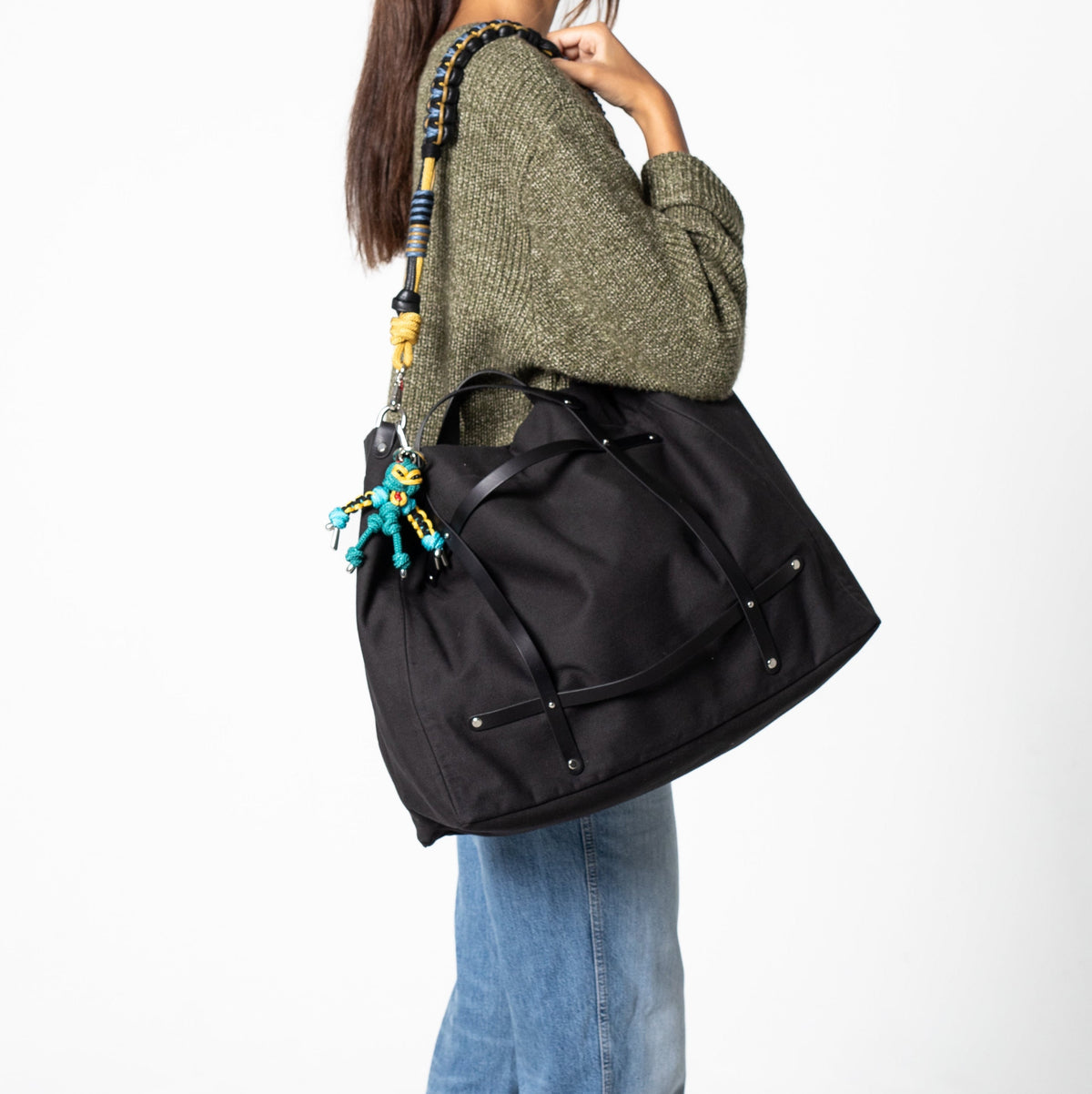 A person wearing a green knit sweater and blue jeans carries a large black duffel bag with a colorful beaded shoulder strap and tassel charm against a plain white background.