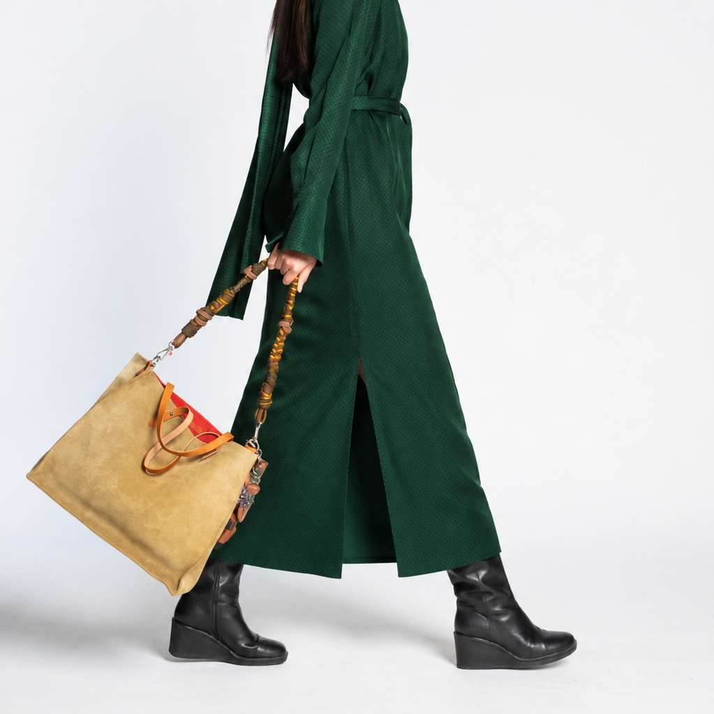 A model stands against a light gray studio background wearing a dark green long-sleeve midi dress with a waist tie, holding a tan tote bag with brown leather handles and tassels.