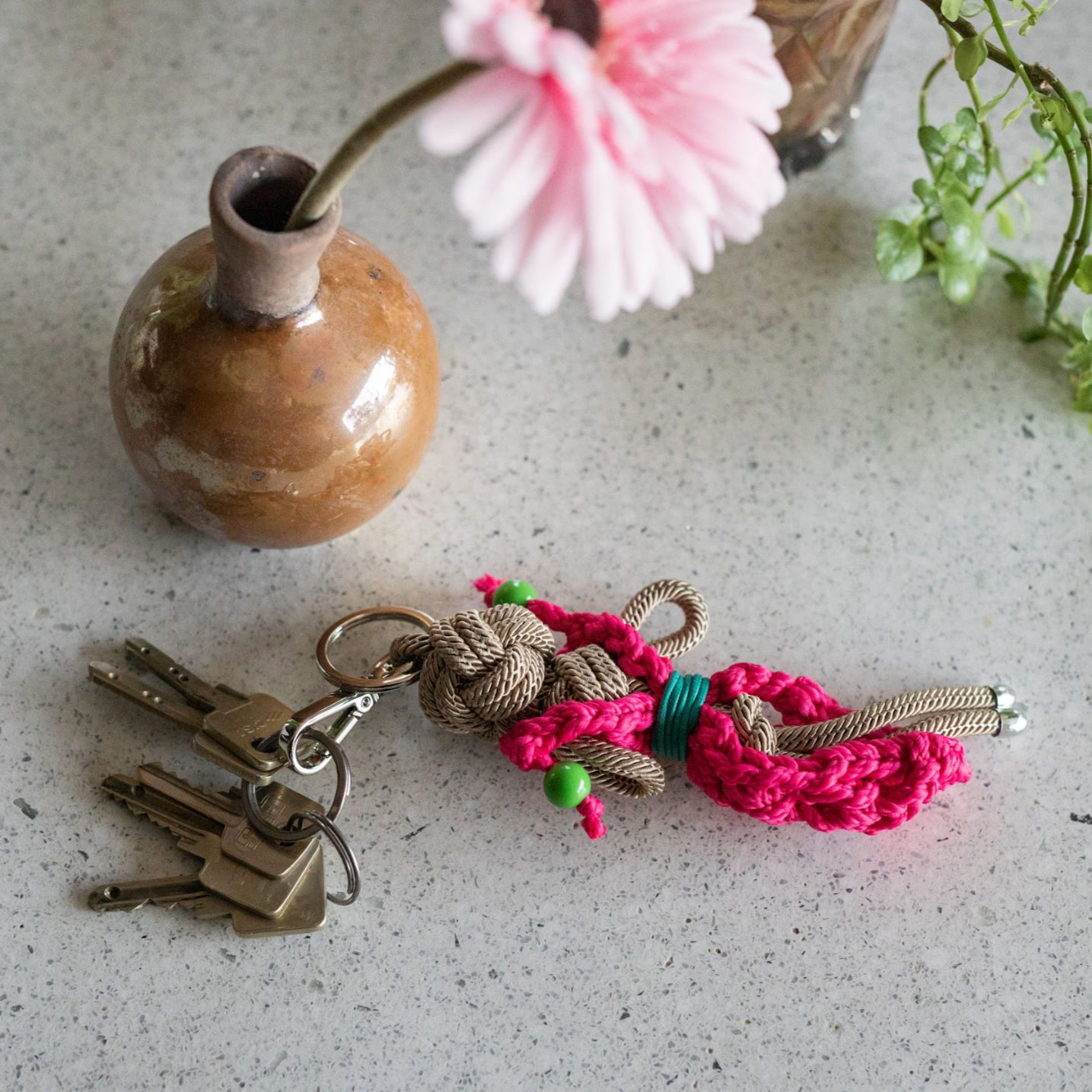 Lucinda bag charm with keys attached and laid on a marble surface near a vase with a singular pink flower in it.