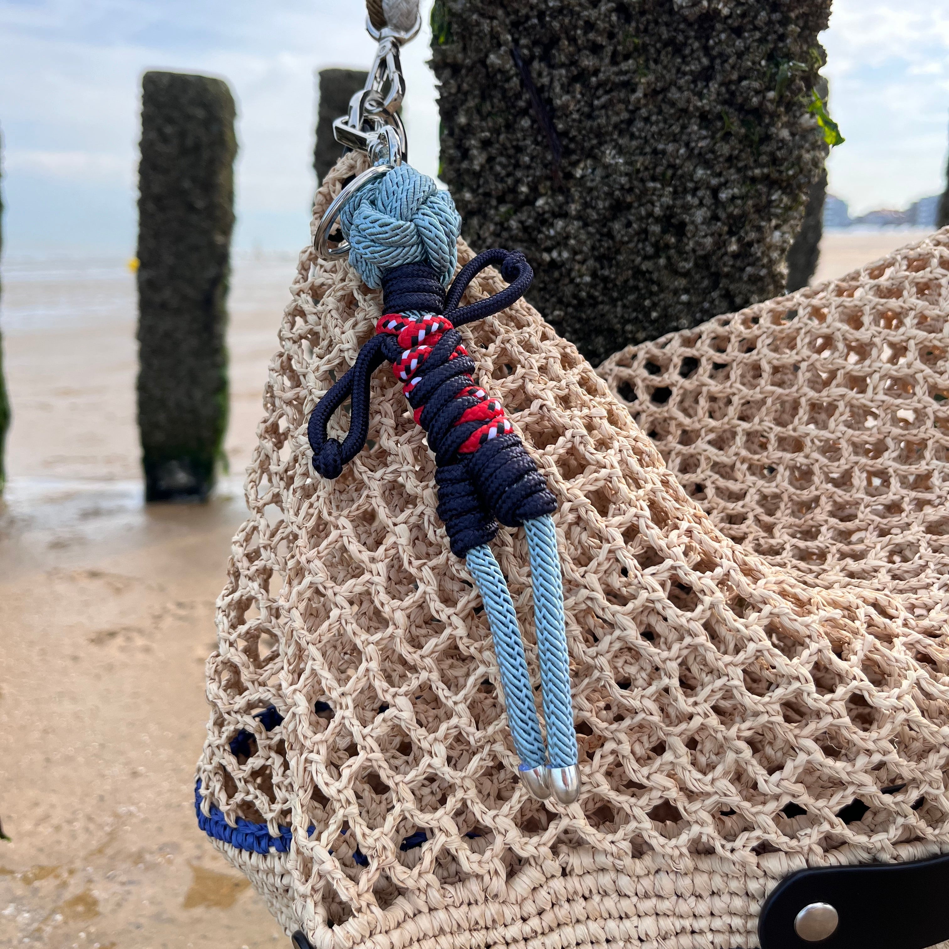 A close-up of a beige raffia tote bag with the Gwen bag charm attached, photographed outdoors near a beach and water in the background.