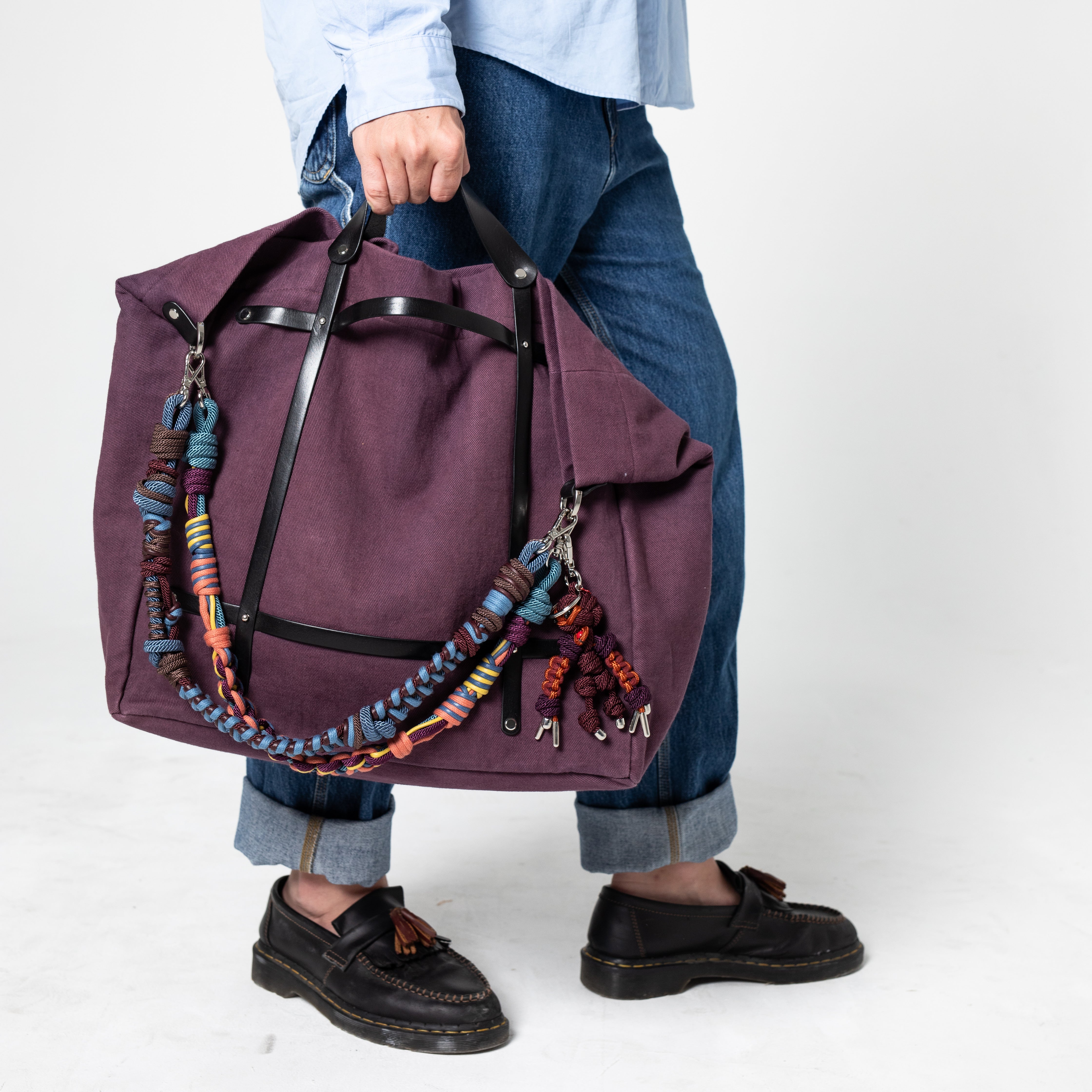 Model holding an aubergine weekend or duffle bag with colorful macramé shoulder straps.