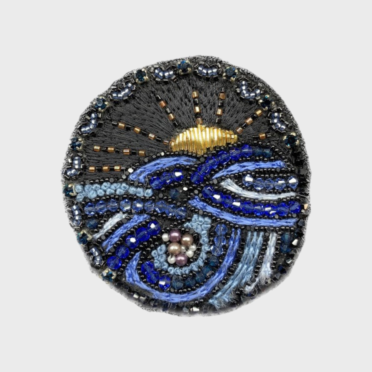 An embroidered brooch with a drawing of a sun setting in the sea with black, blue and yellow details. 