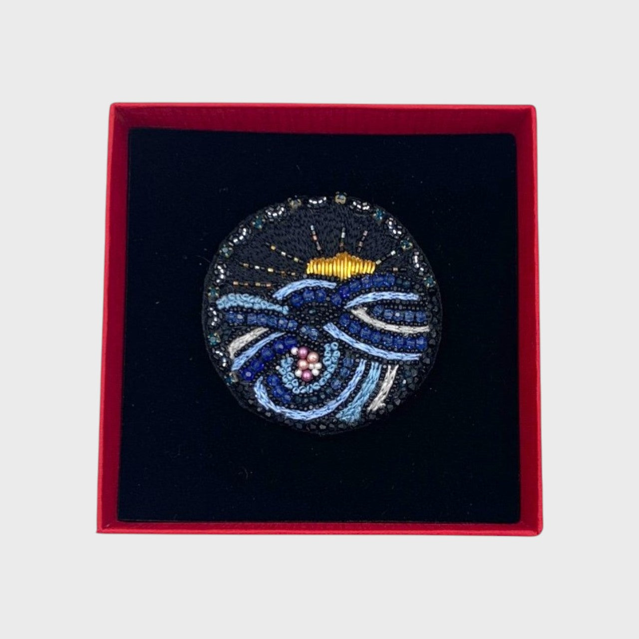 An embroidered brooch representing the sun setting down in the sea in a box. 