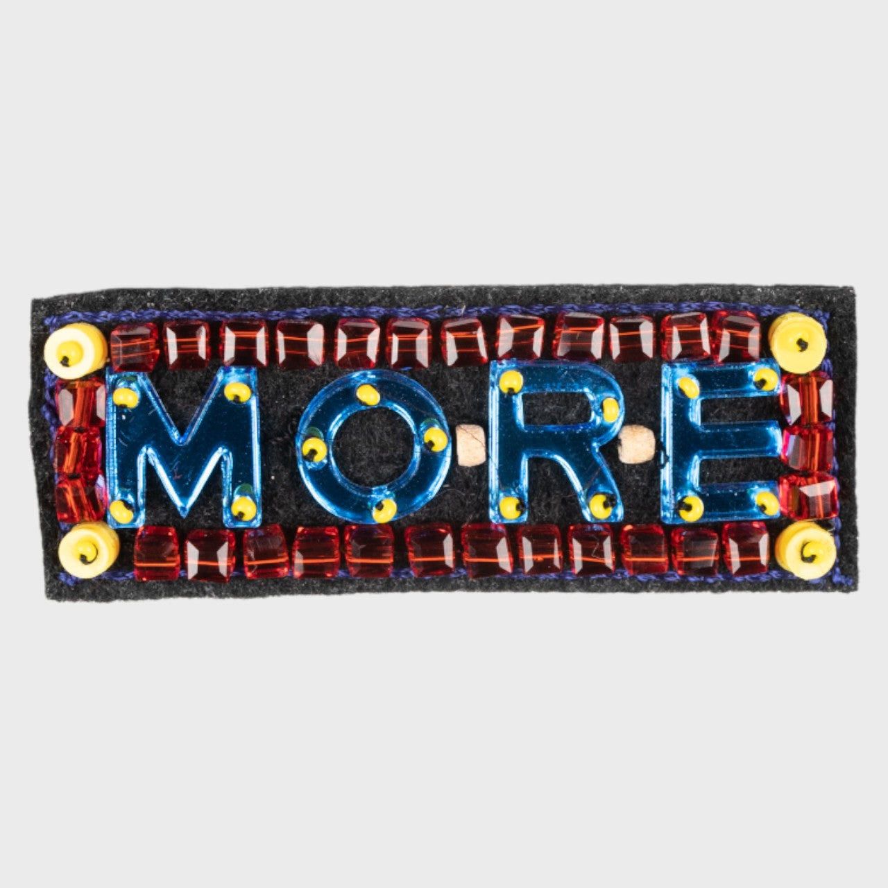 An embroidered brooch with blue MORE with red details. 