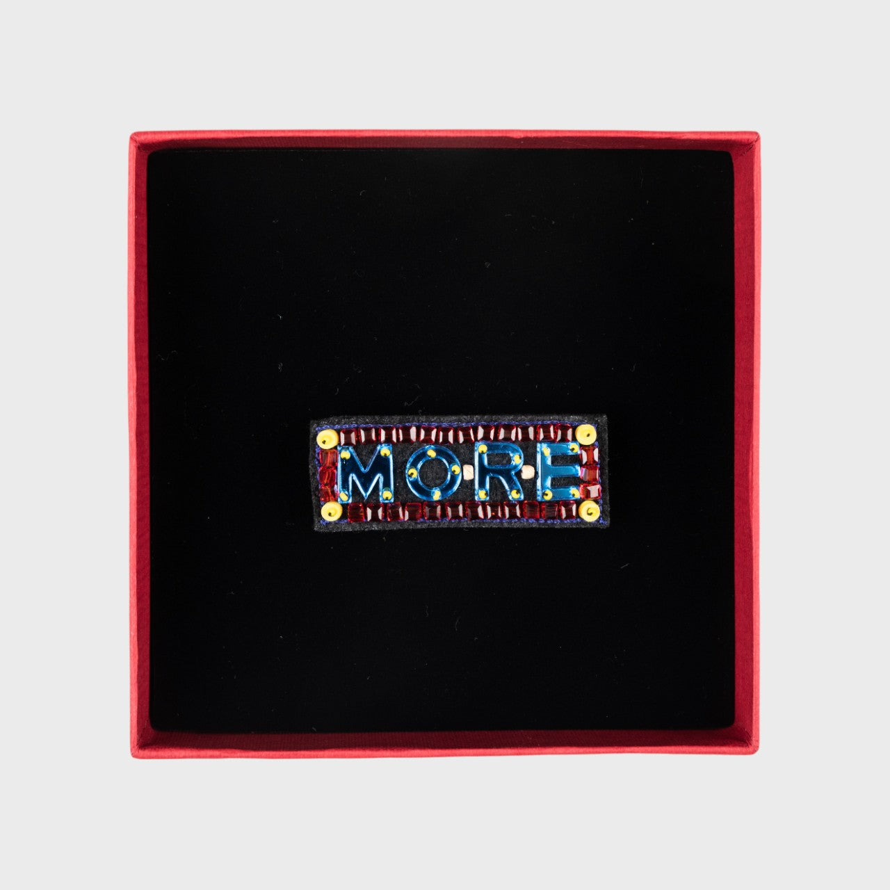 An embroidered brooch with written More in blue in a box. 