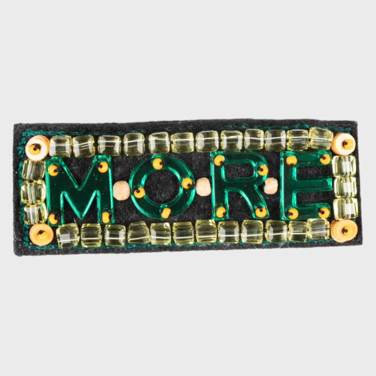 An embroidered green brooch with written MORE with yellow details. 