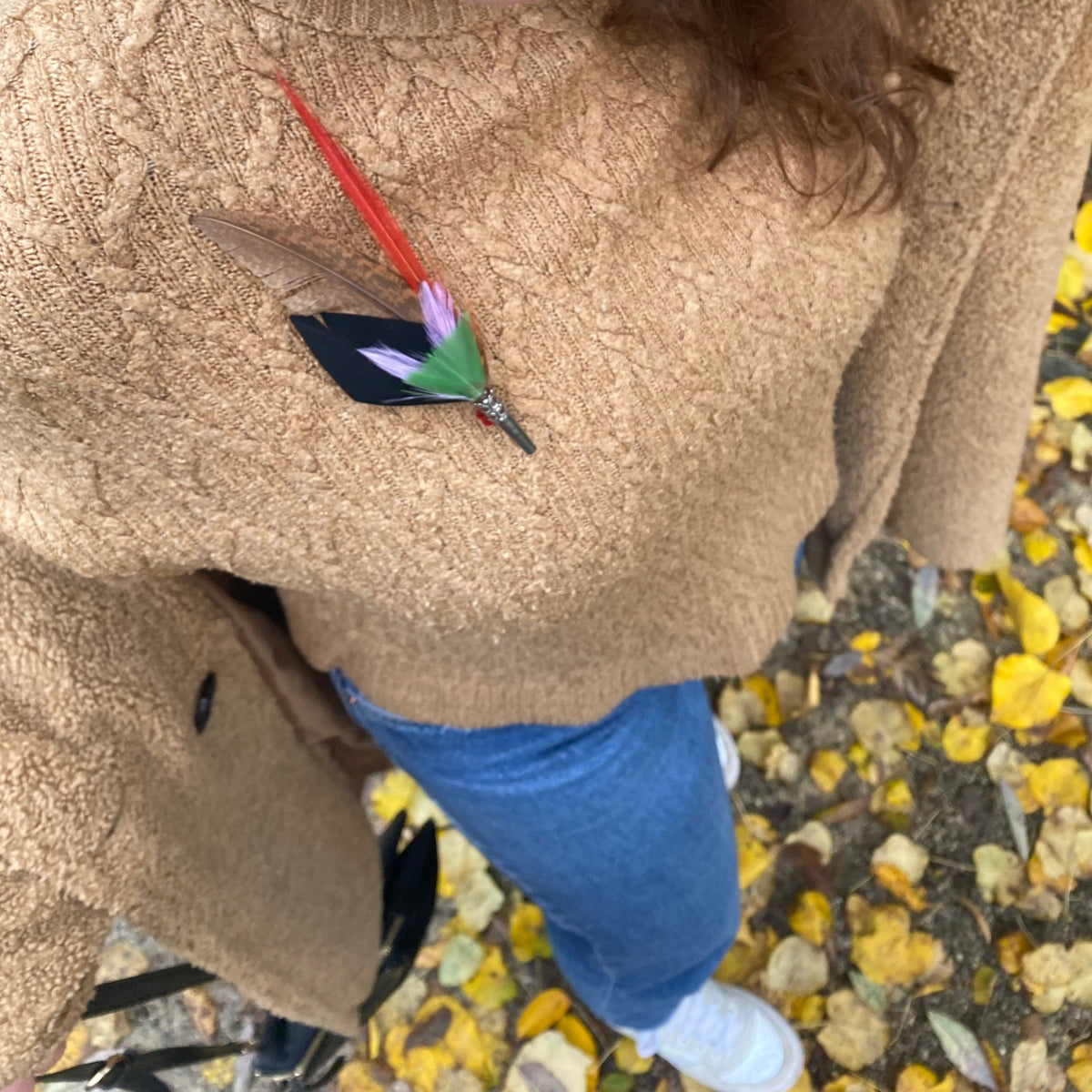a woman wearing a brown wool sweater with a feather brooch attached to it, against a backdrop of autumn leaves.