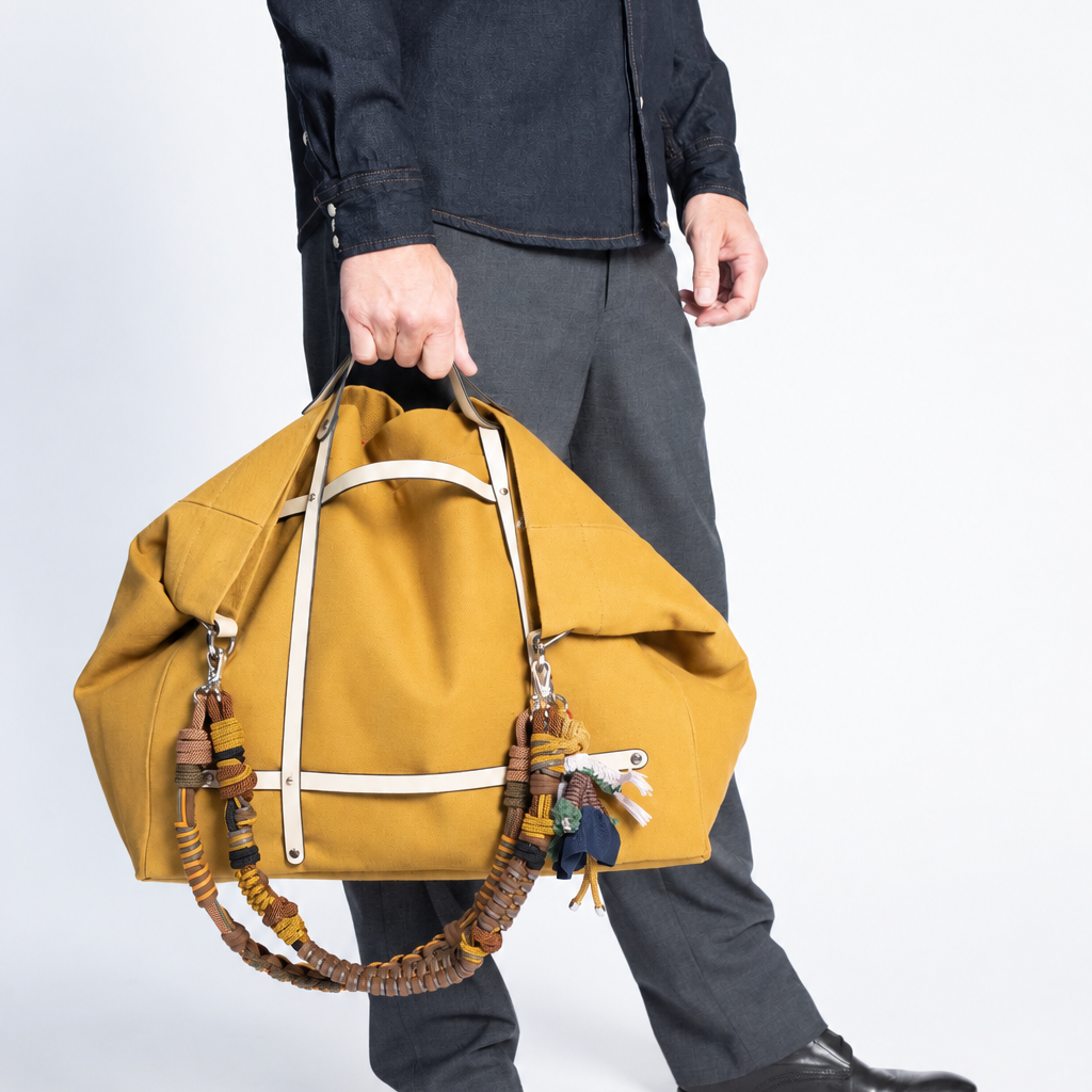 A mustard yellow canvas duffel bag with cream leather straps and a braided brown woven strap detail, held by a person. 