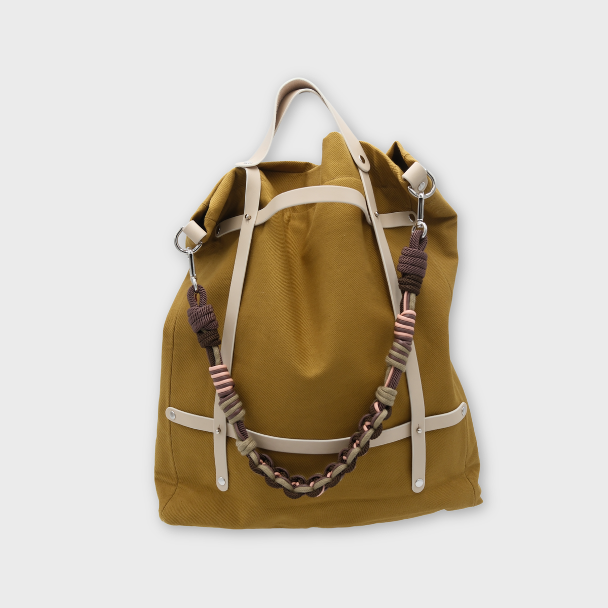 A mustard yellow canvas tote bag with cream-colored leather straps and a chunky woven macrame shoulder strap featuring purple and striped accents, shown against a light gray background.