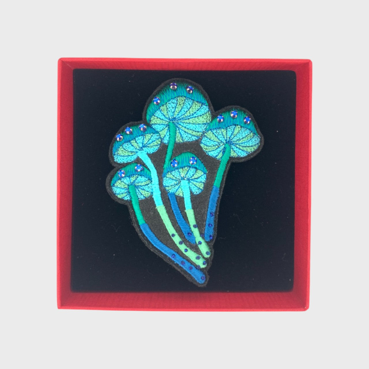 An embroidered brooch representing a bucket of blue mushrooms in a box. 