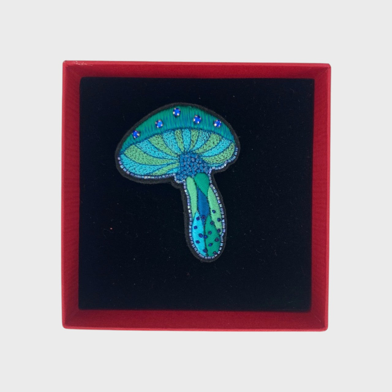 An embroidered brooch representing a blue mushroom in a box. 