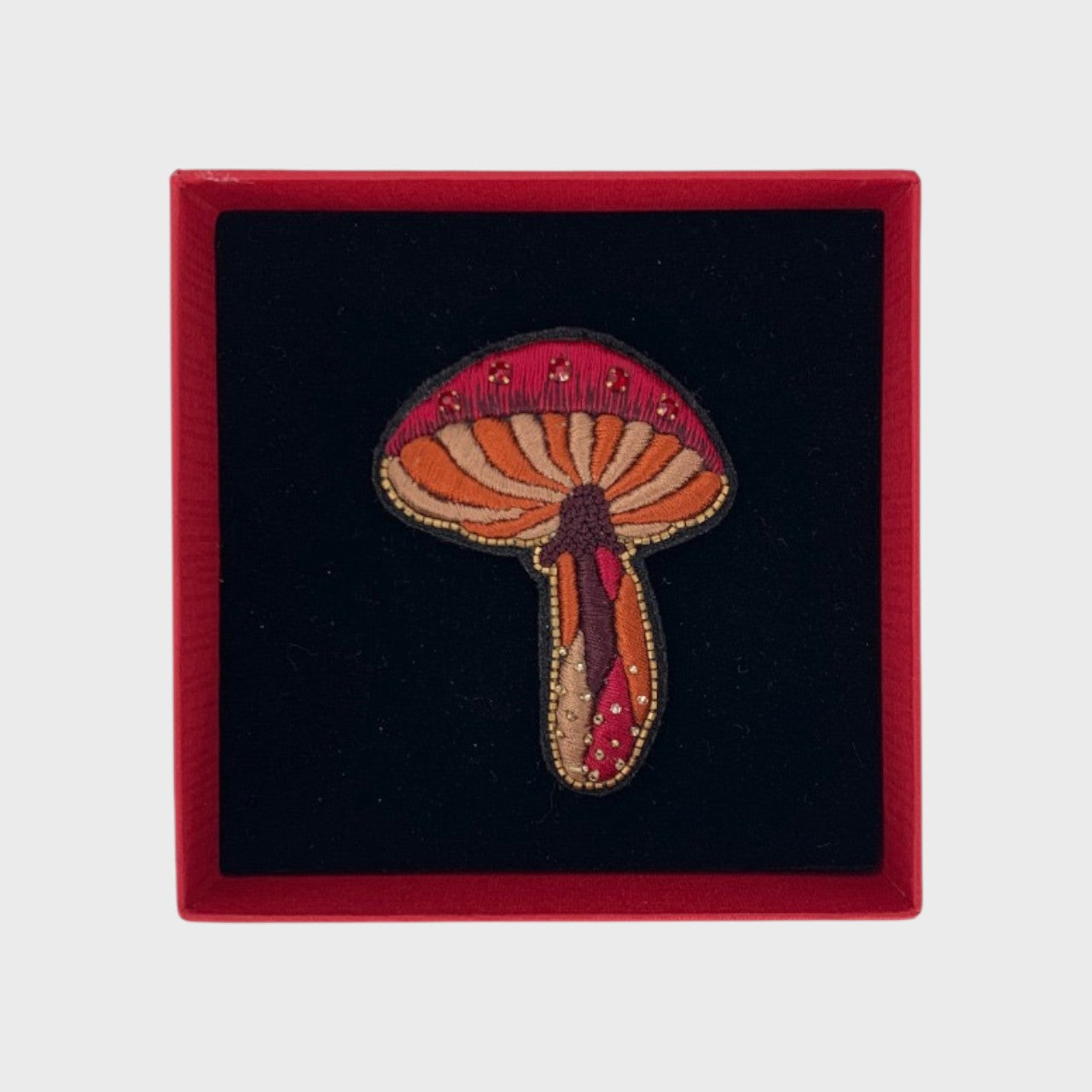 An embroidered brooch representing a red mushroom in a box. 