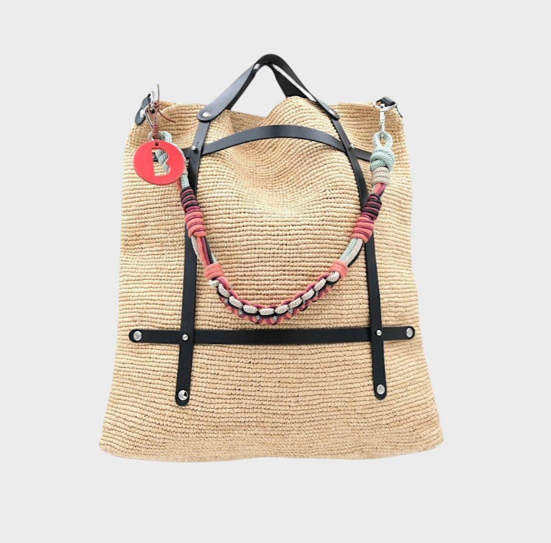 A raffia tote bag in a natural color, featuring black leather straps and a decorative beaded shoulder strap with blue, pink and burgundy details. 