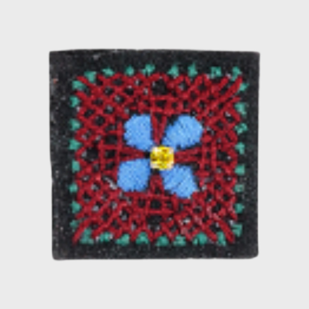 An embroidered brooch representing a square with red and green details and a blue flower in the middle. 
