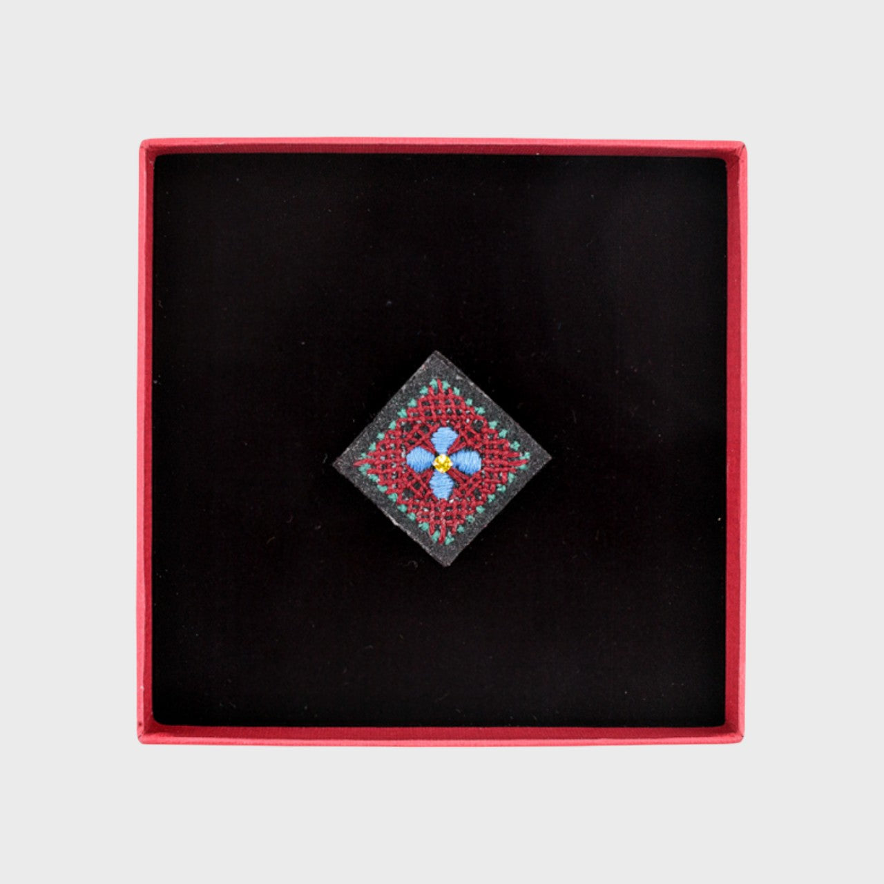 An embroidered brooch representing a square with a blue flower in the middle in a box. 