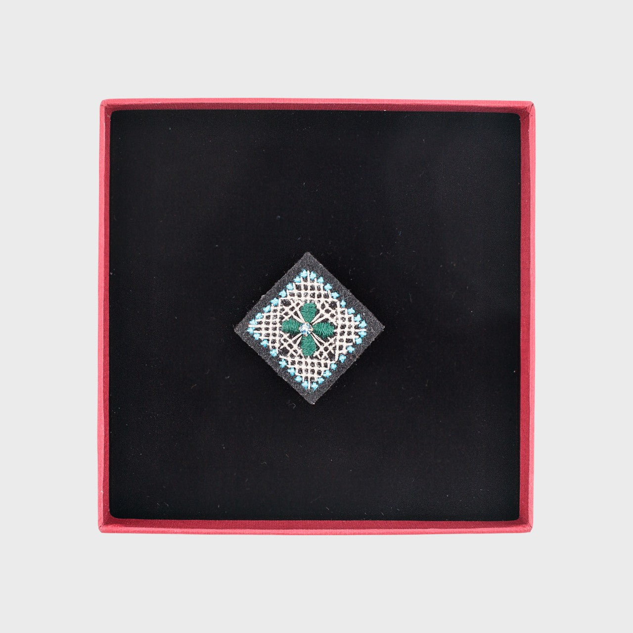 An embroidered brooch representing a square with a green flower in the middle in a box. 