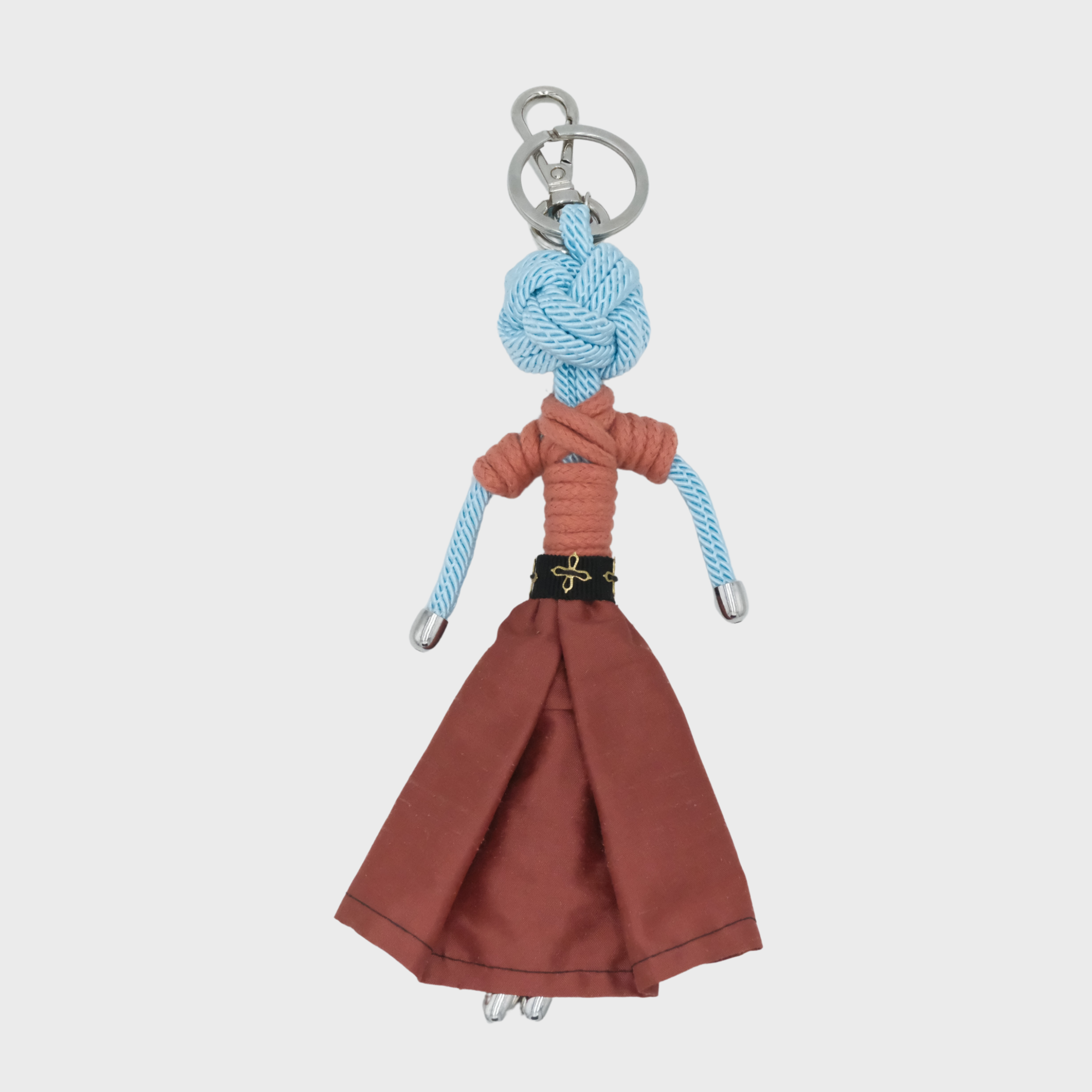 A handmade macramé doll keychain featuring a light blue braided rope head and arms, a rust-orange short-sleeve top, a rust-brown skirt, and a black belt with a gold detailed buckle, attached to a silver metal key ring.