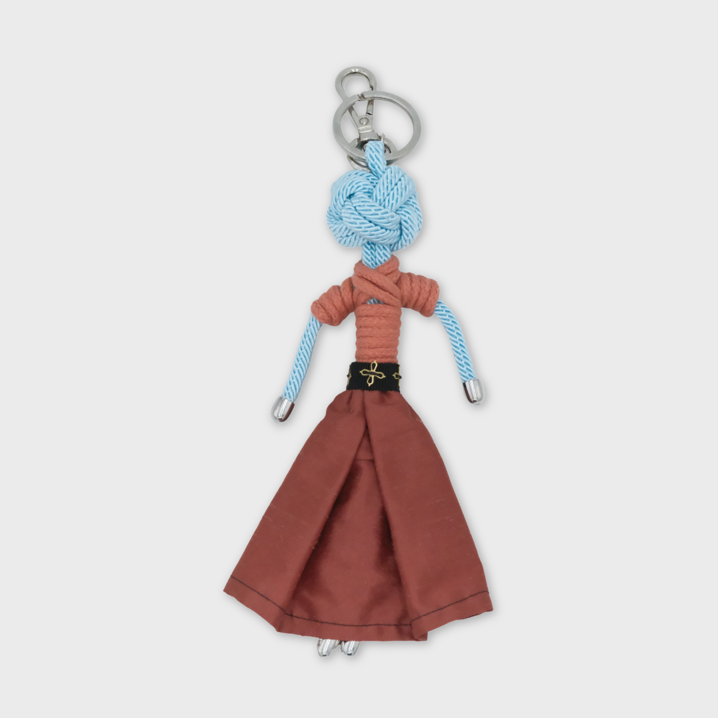 A handmade macramé doll keychain featuring a light blue braided rope head and arms, a rust-orange short-sleeve top, a rust-brown skirt, and a black belt with a gold detailed buckle, attached to a silver metal key ring.
