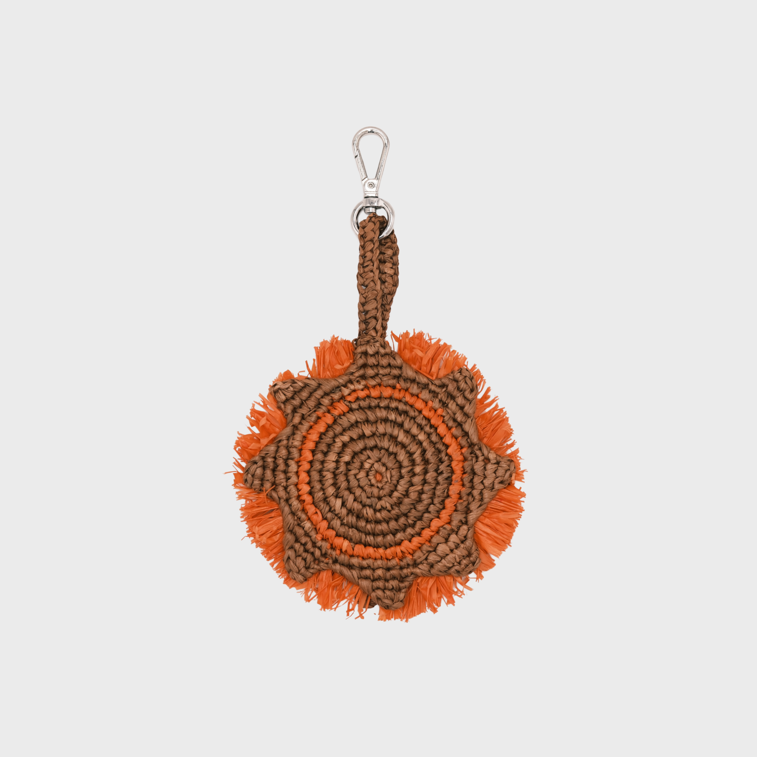 A circular raffia keychain with a spiral pattern in brown and orange, representing a sun and featuring fringe details and a silver metal lobster clasp at the top.