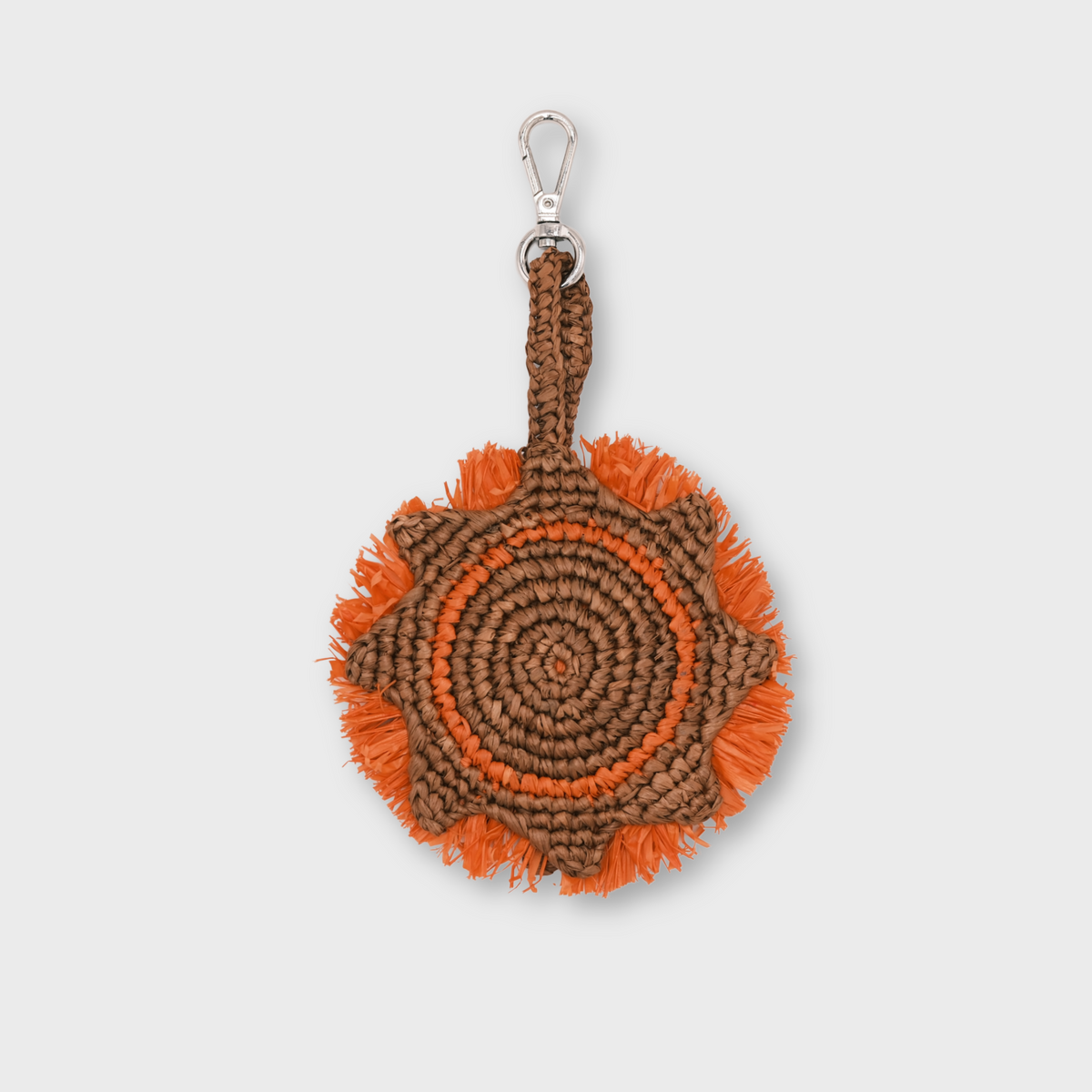 A circular raffia keychain with a spiral pattern in brown and orange, representing a sun and featuring fringe details and a silver metal lobster clasp at the top.