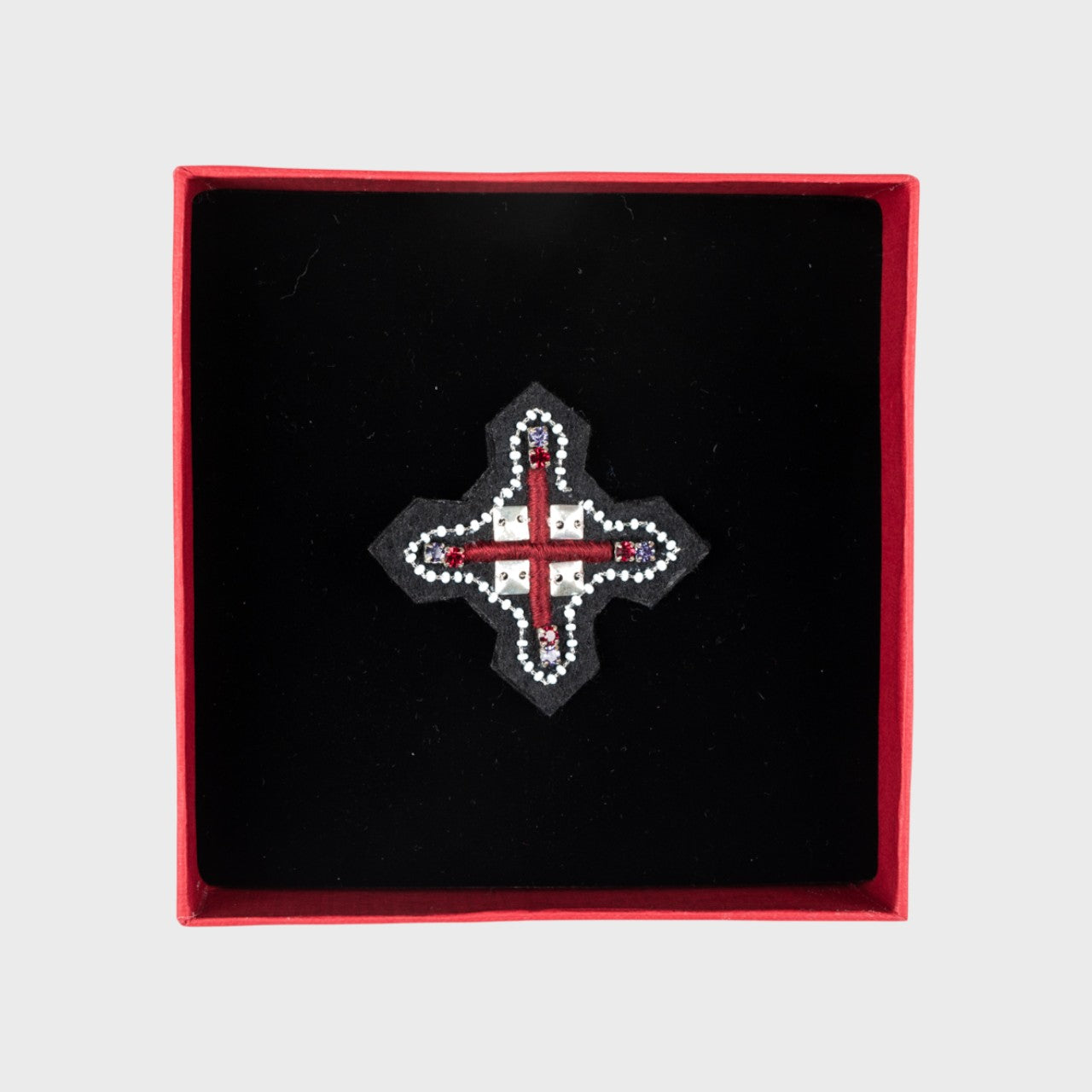 An embroidered brooch representing a red cross with white details in a box. 
