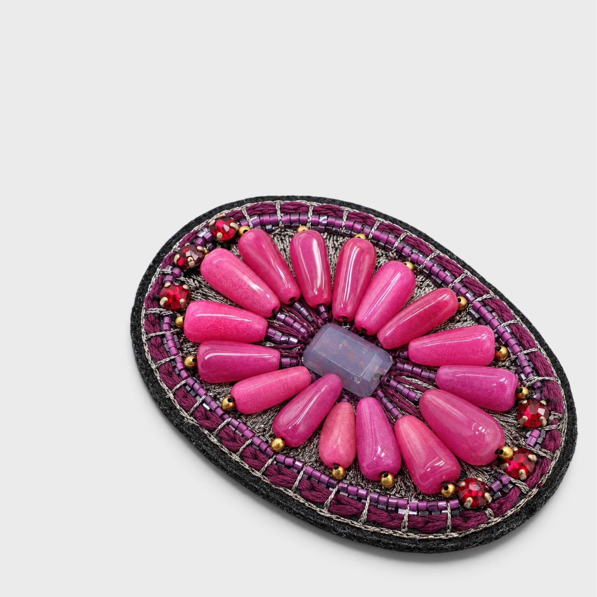 An oval beaded brooch featuring a central purple rectangular stone surrounded by radiating pink and magenta beads, with a dark purple beaded border and small red and yellow accents, set against a light gray background.