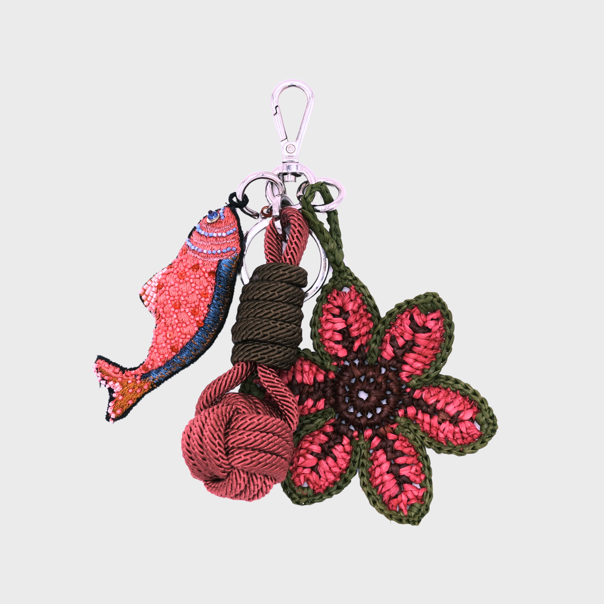 A handmade macramé and raffia keychain featuring a pink and blue fish charm, a knotted prink and green ball, and a pink and green raffia flower, attached to a silver clasp.