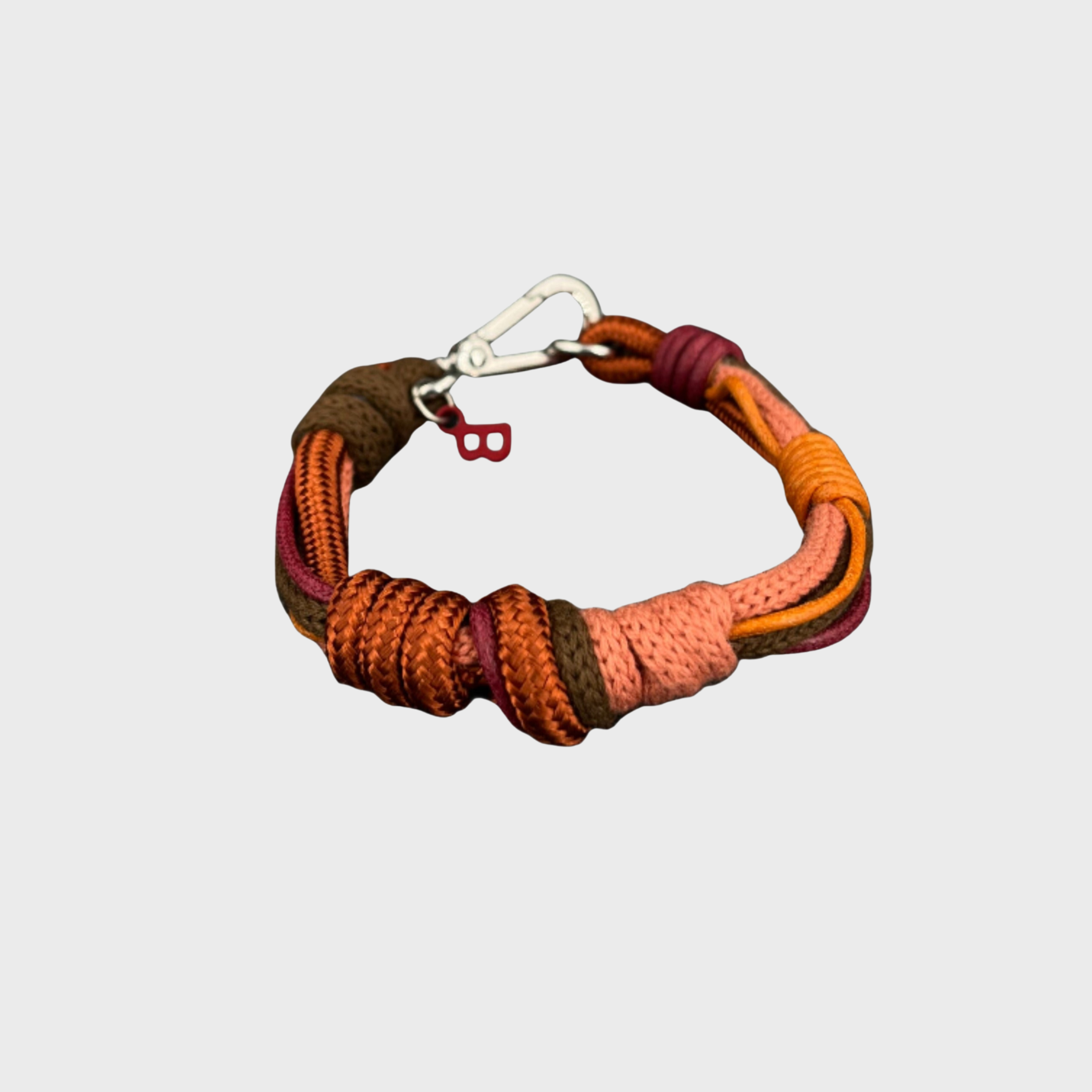 A macramé bracelet with knotted details, featuring shades of pink and brown, finished with a silver-tone metal clasp, set on a light gray background.